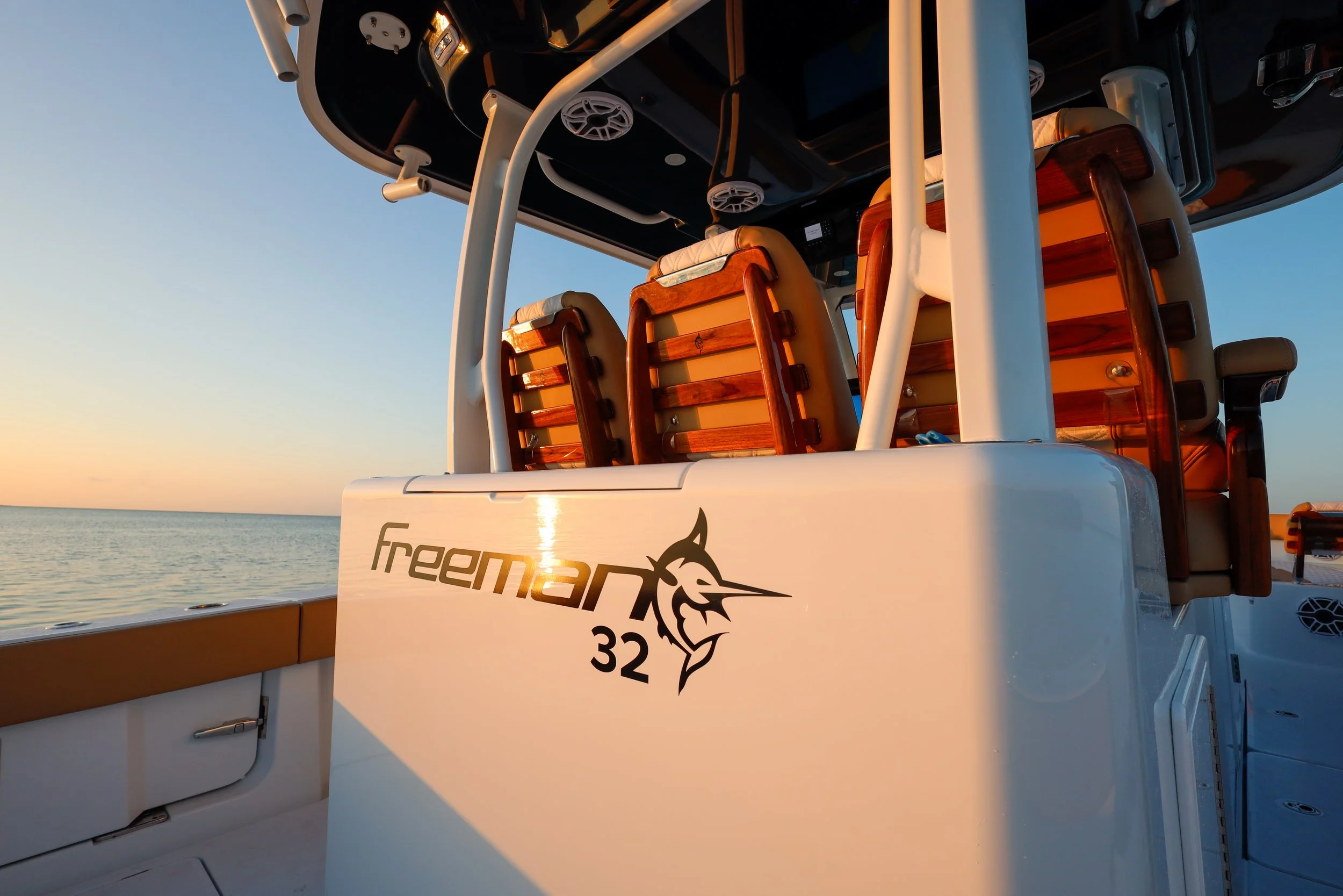 The back of a white Freeman 32 boat with three wooden chairs and a part of the ocean visible during sunset.