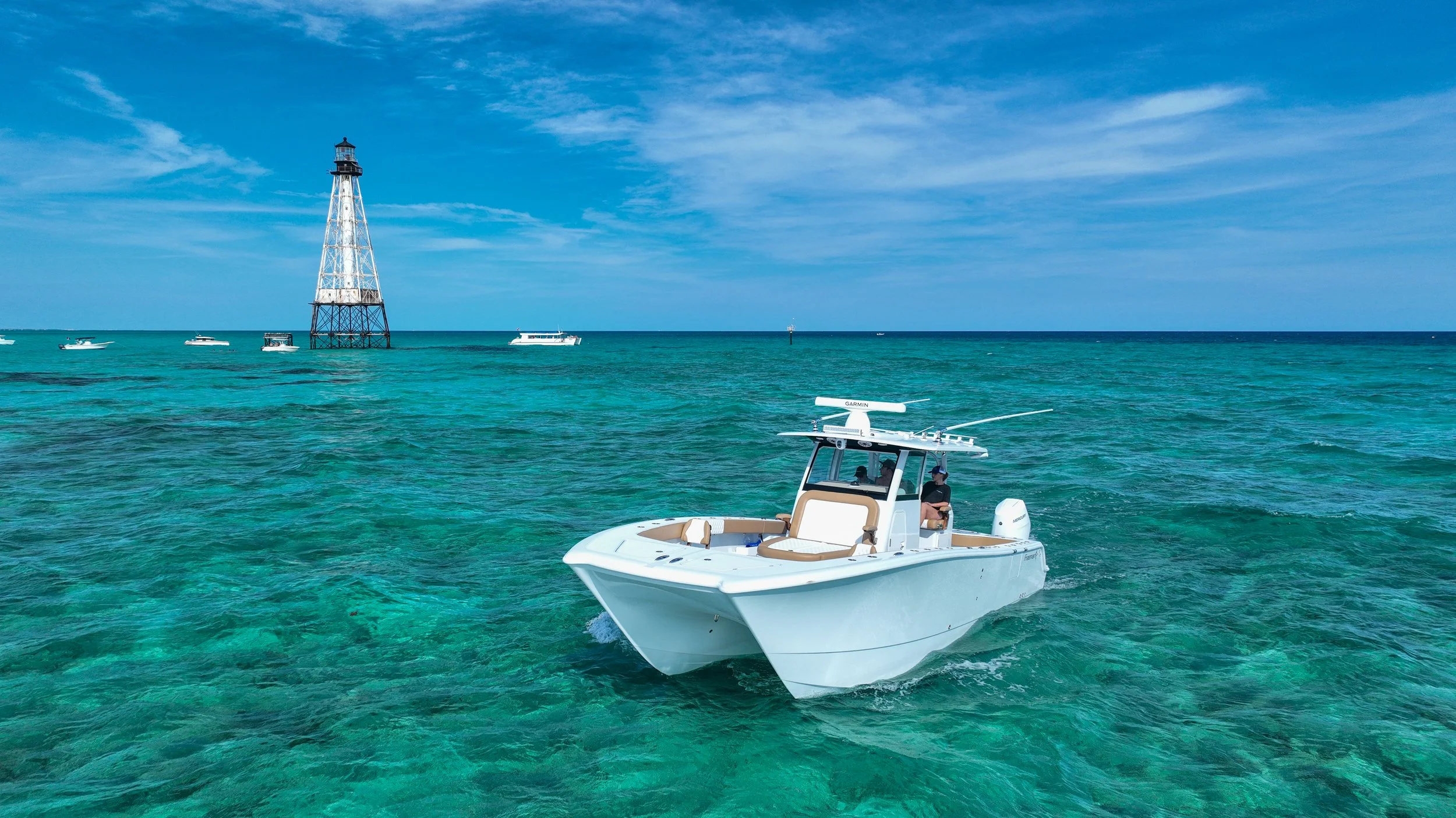 A white boat with tan and white seats navigating turquoise waters near a lighthouse and several other boats under a partly cloudy sky.