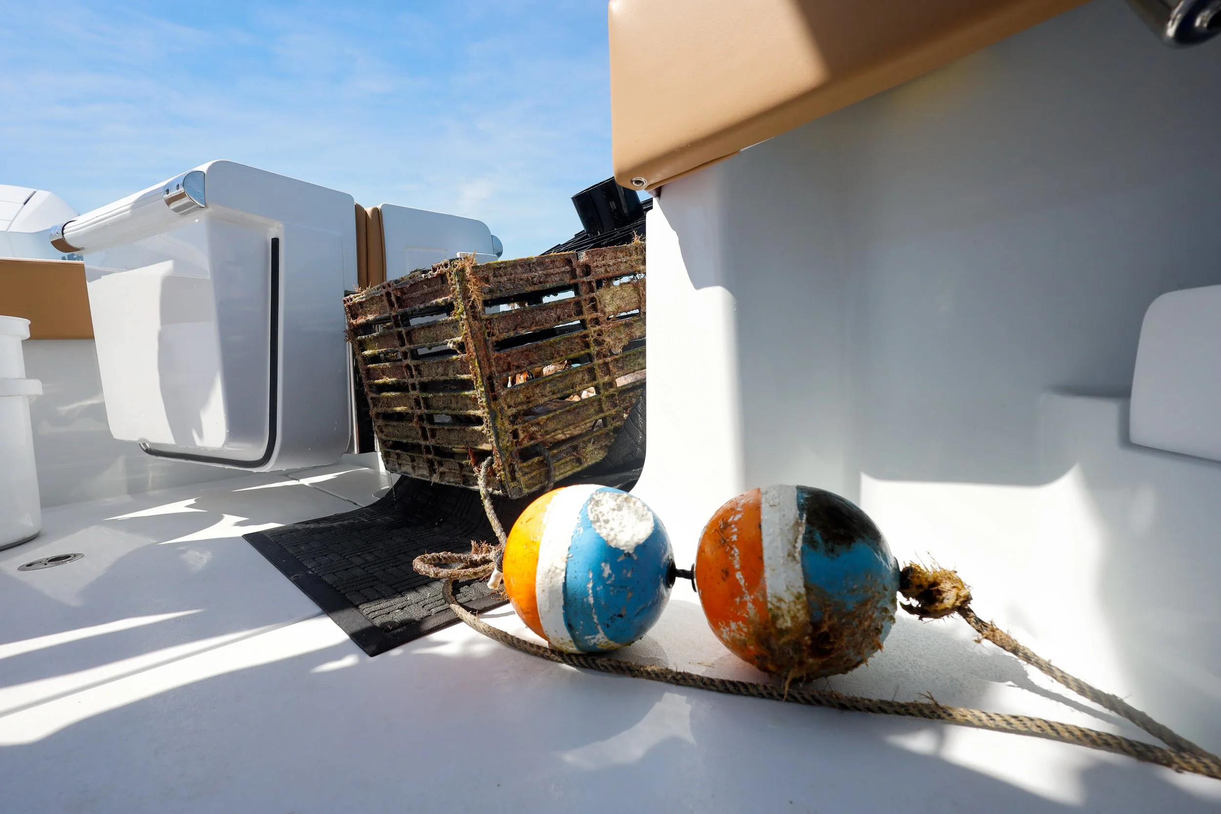 Close-up of weathered orange, white, and blue boat fenders, a rusty metal crate, and parts of the boat's deck with white surfaces and some equipment.