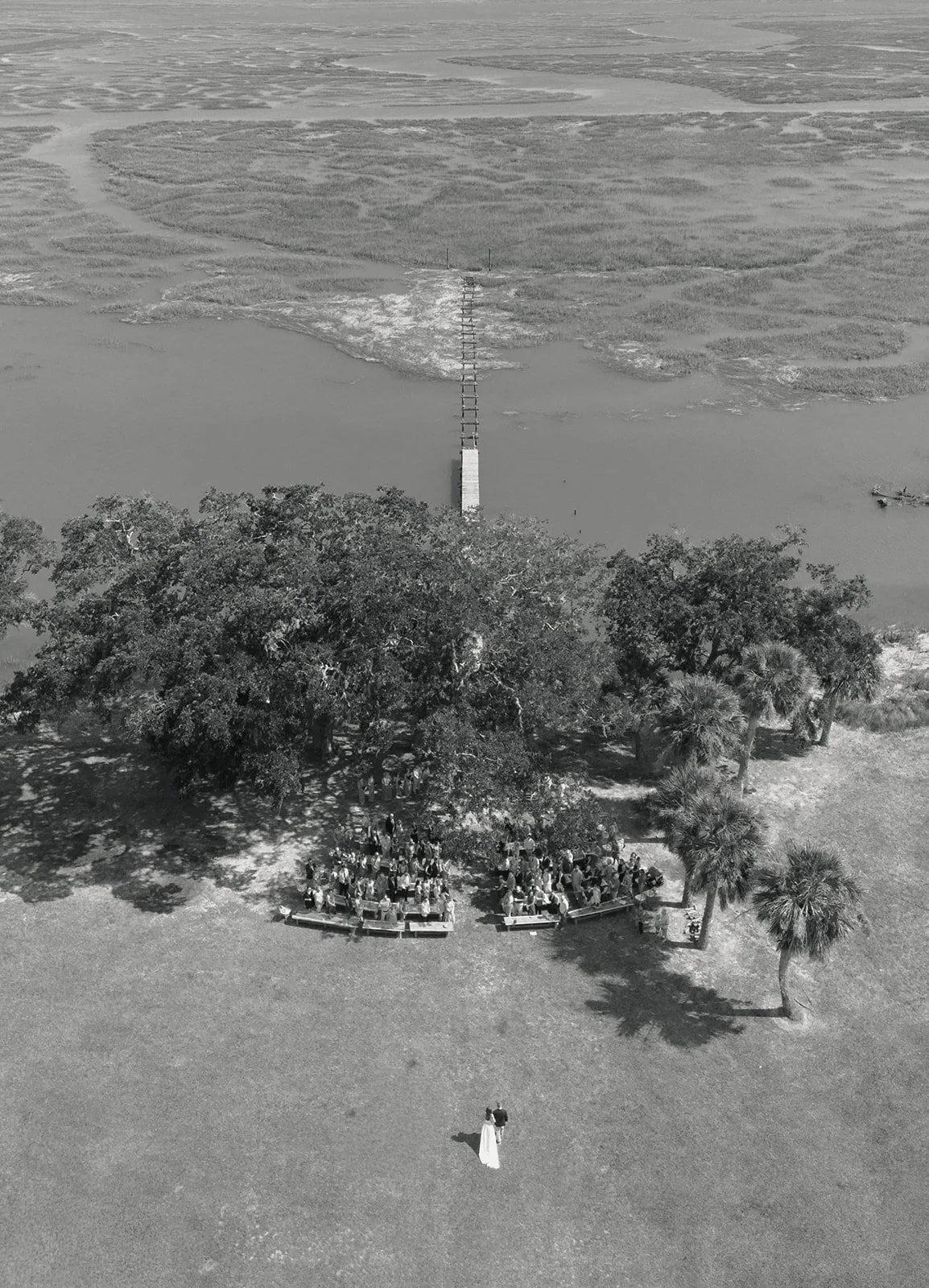 A black and white aerial photo of a wedding ceremony taking place outdoors by a lakeside, with a bride and groom standing in front of a group of guests under large trees, and a dock extending into the water.