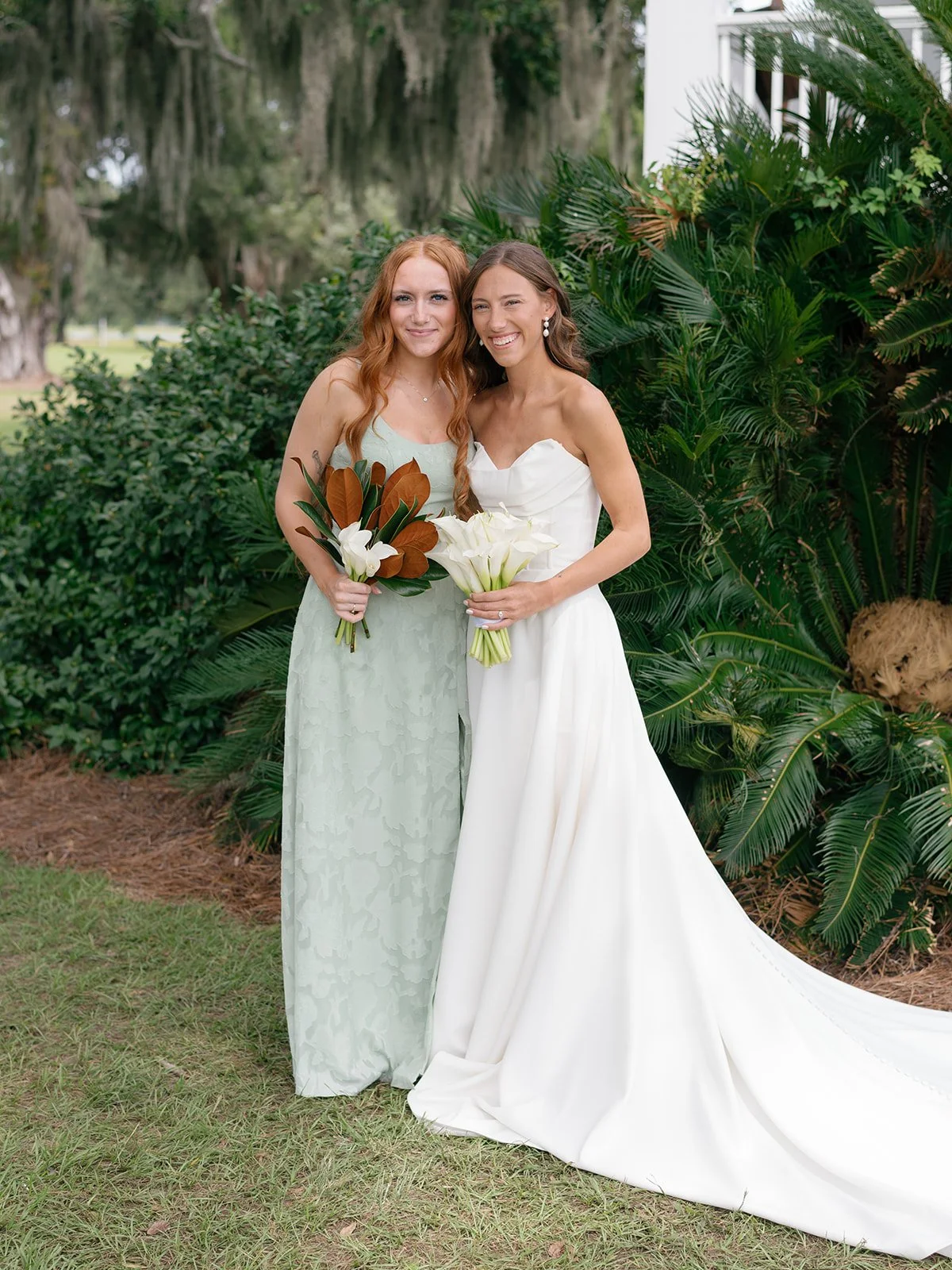 Two women, one in a white wedding dress and the other in a light green dress, standing outdoors with greenery in the background. They are smiling and holding bouquets of white flowers.