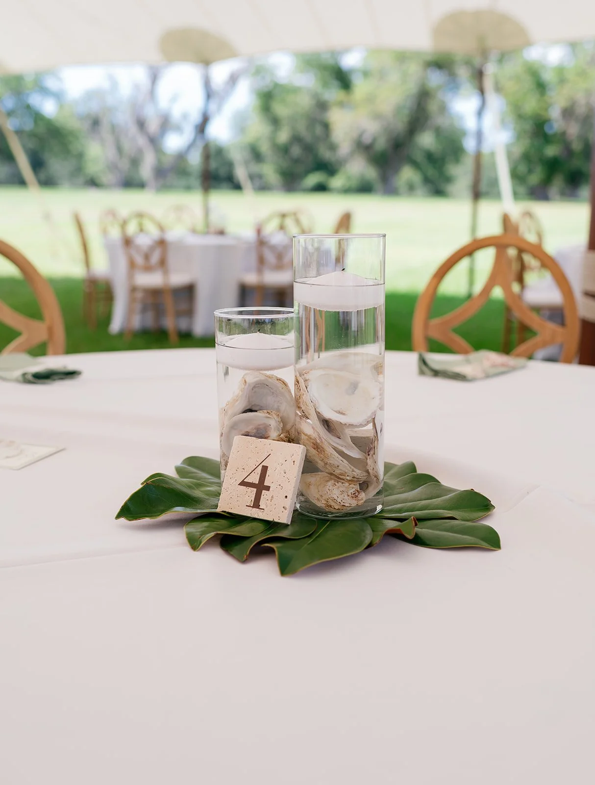 Table with glass vases filled with water and seashells, numbered 4, on a large green leaf at an outdoor event with round tables, chairs, and umbrellas in the background.