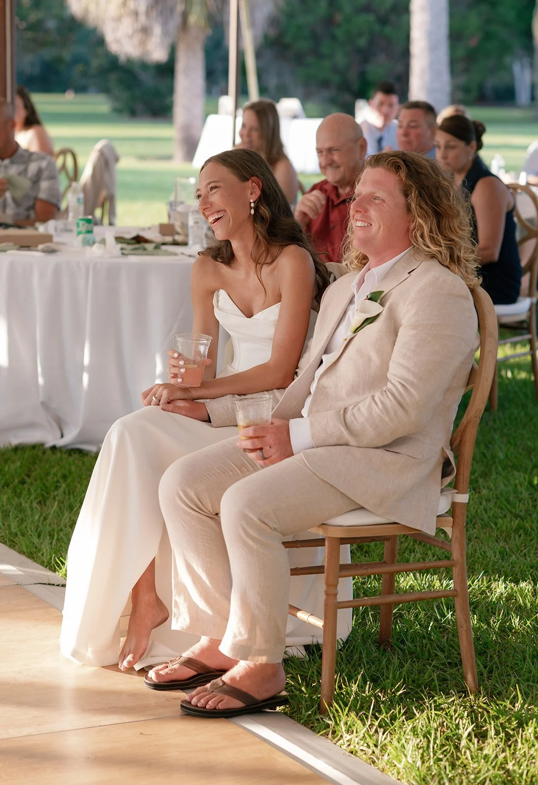 A bride and groom sitting on chairs at a wedding reception, smiling and enjoying drinks, with guests seated at tables in the background outdoors.