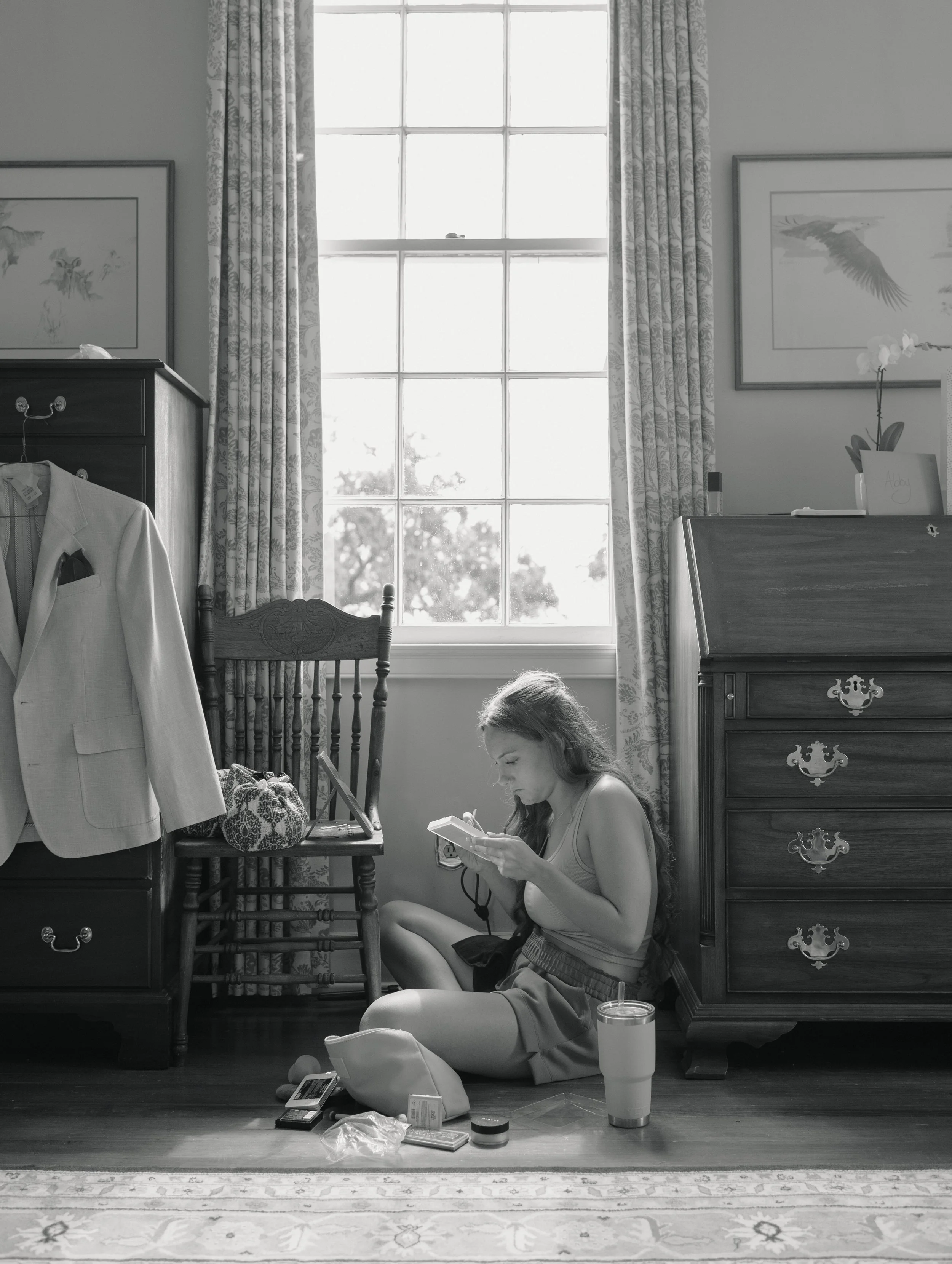A woman sitting on the floor in front of a dresser, looking at her phone, with various items on the floor including a fast food drink, containers, and a phone. The room has a large window with patterned curtains, a wooden chair, and framed artwork on