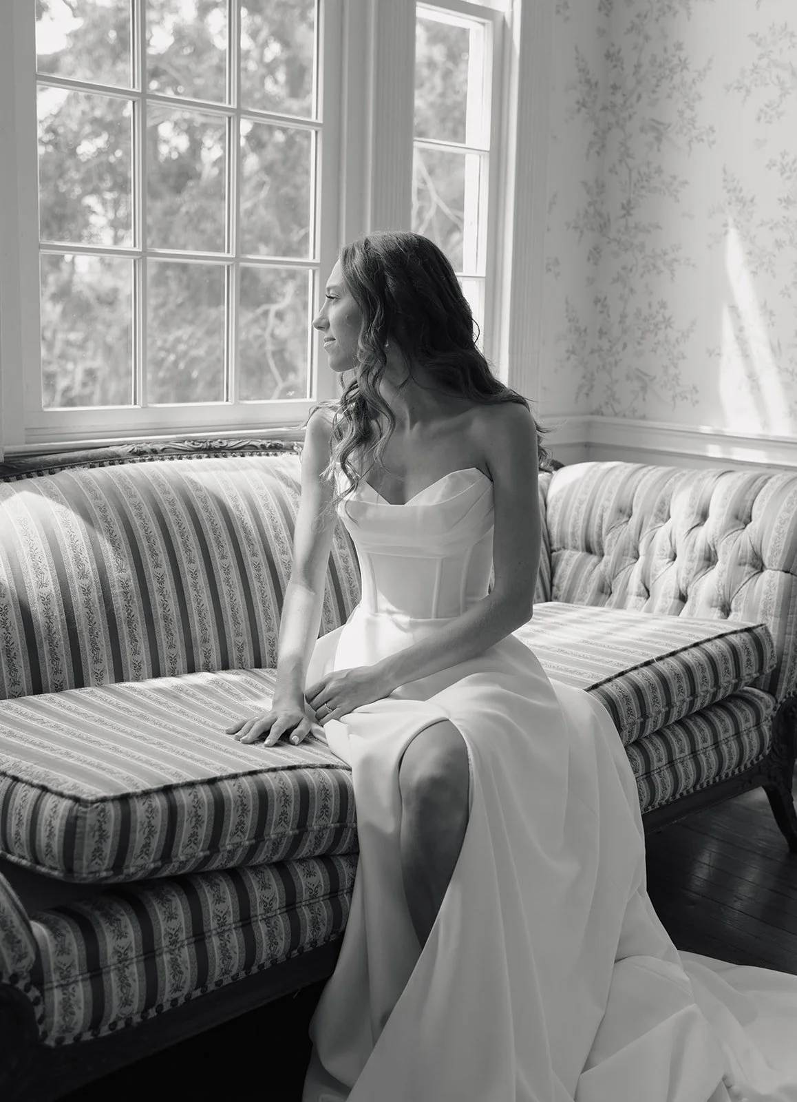 A woman in an elegant strapless wedding gown sitting on a vintage patterned sofa, looking out the window in a room with floral wallpaper.