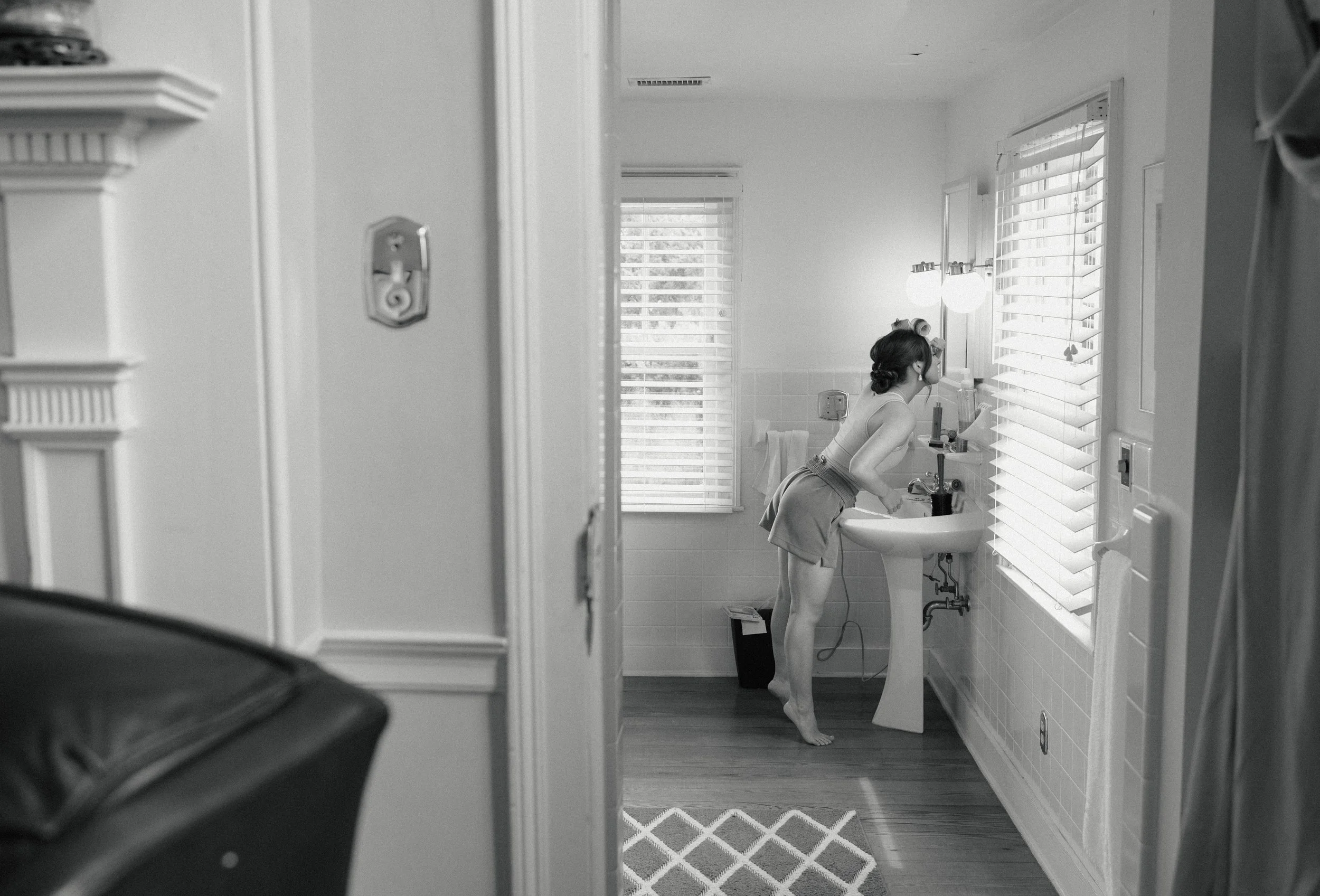 A woman leaning over a bathroom sink, looking into the mirror, with her hair tied back, wearing a sleeveless top and shorts, in a well-lit bathroom with horizontal blinds on the windows.