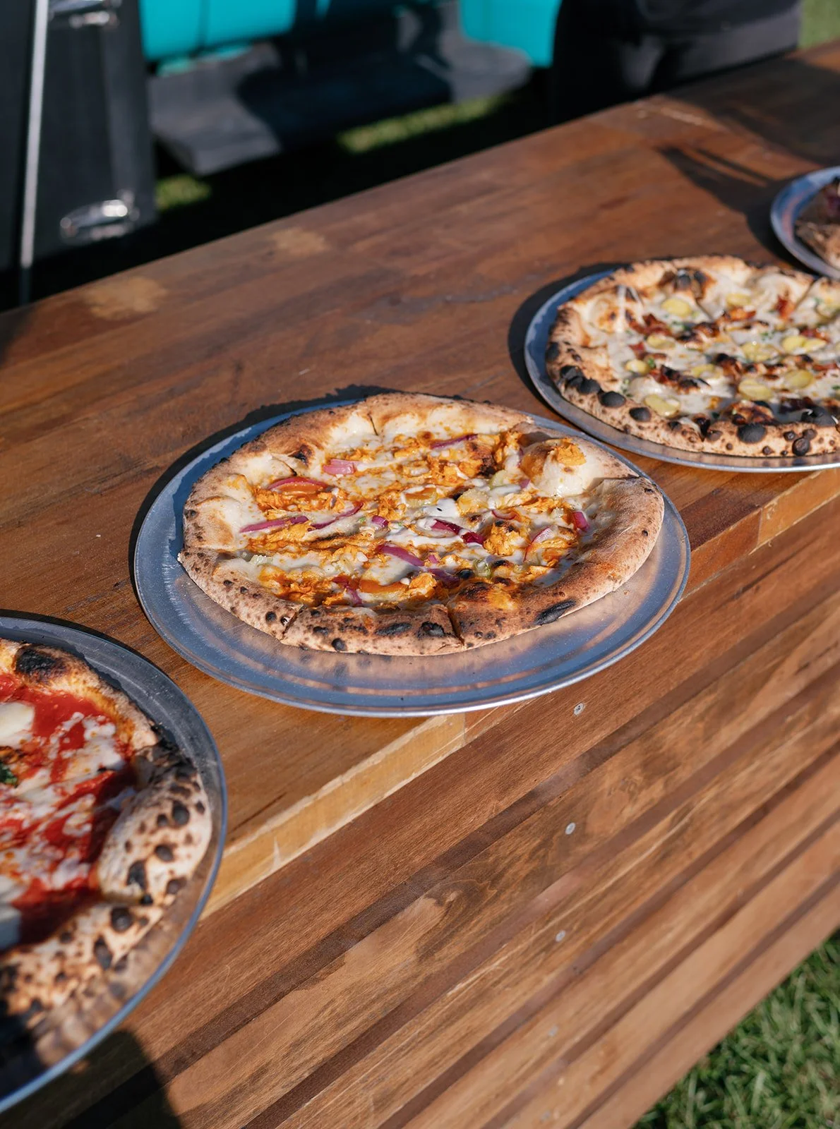 Three pizza pies on clear plastic trays placed on a wooden table outdoors.