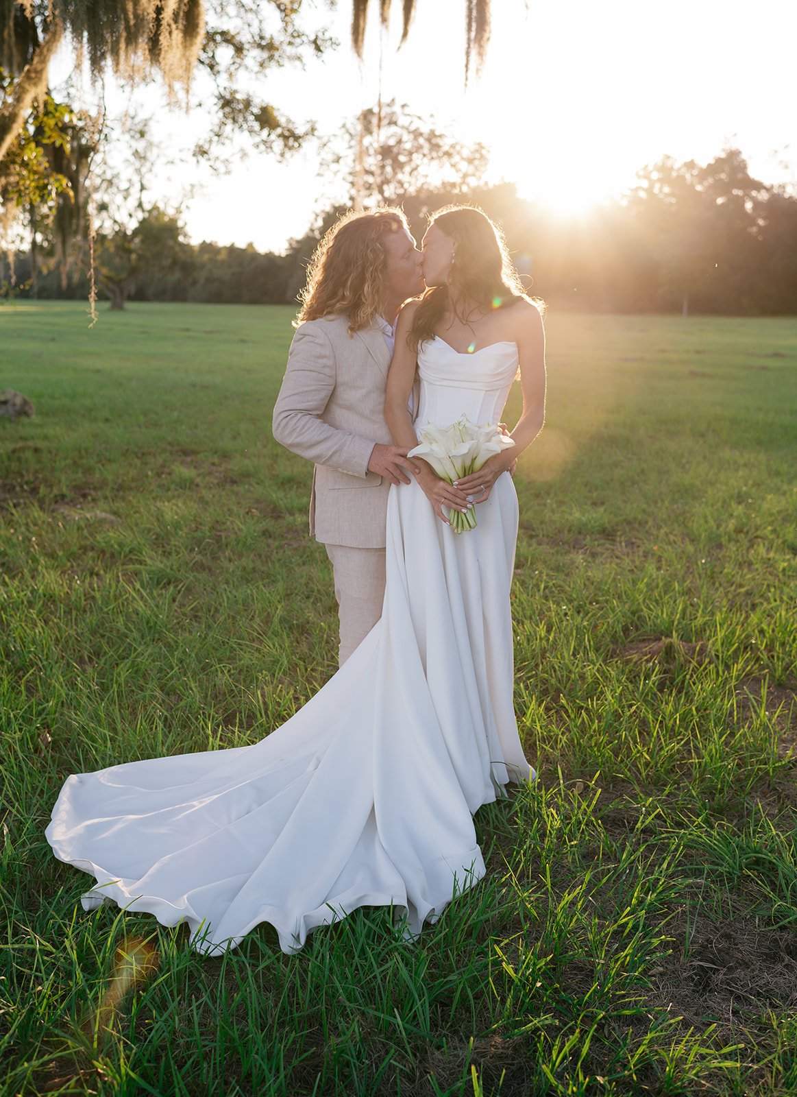 An engaged couple kissing outdoors in a grassy field during sunset, the woman in a white wedding gown holding a bouquet of calla lilies, and the man in a light-colored suit.