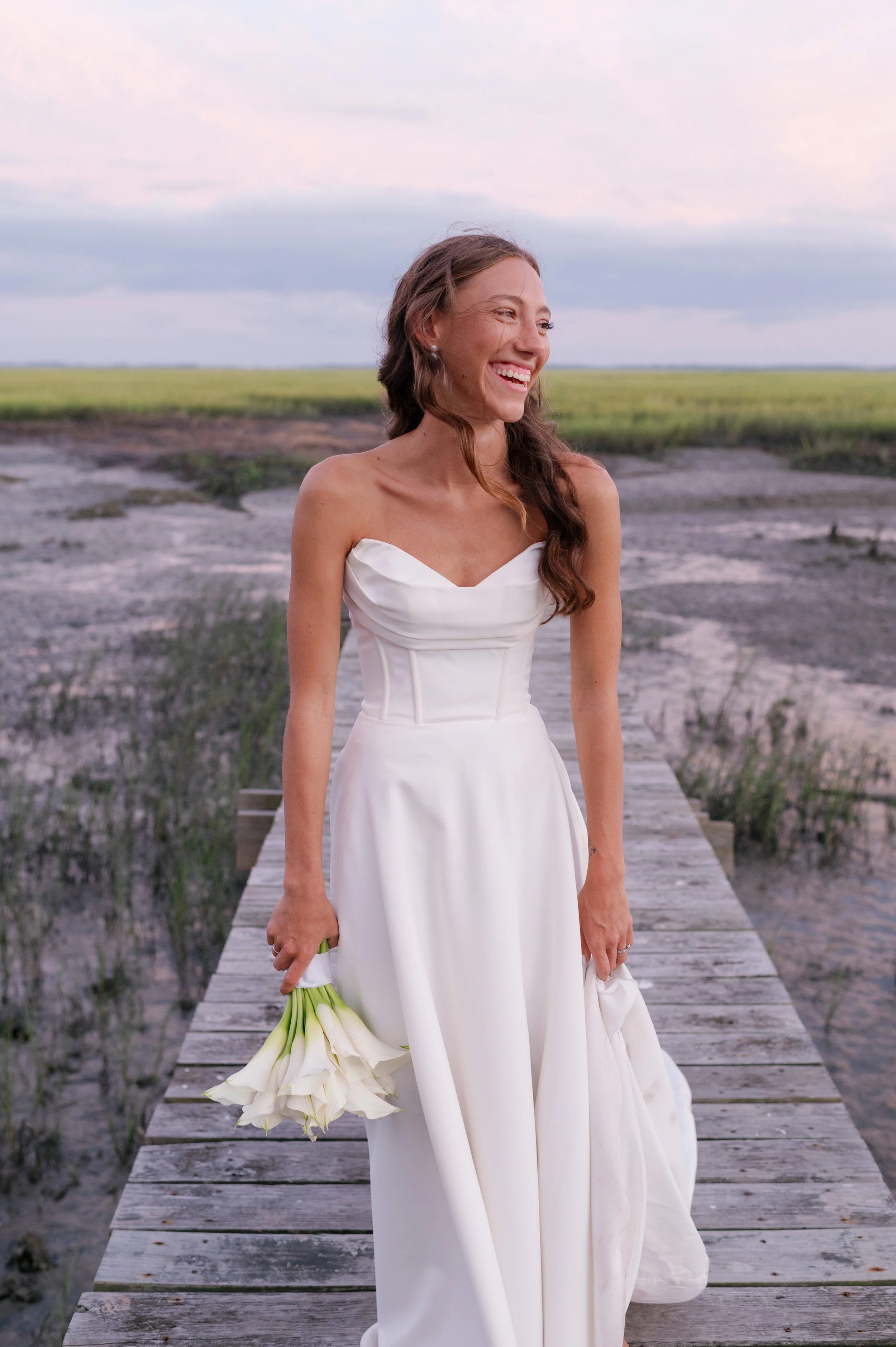 A woman in a white strapless wedding dress stands on a weathered wooden dock holding a bouquet of white calla lilies. She is smiling and looking to her right, with a landscape of flat grassy fields and a cloudy sky in the background.