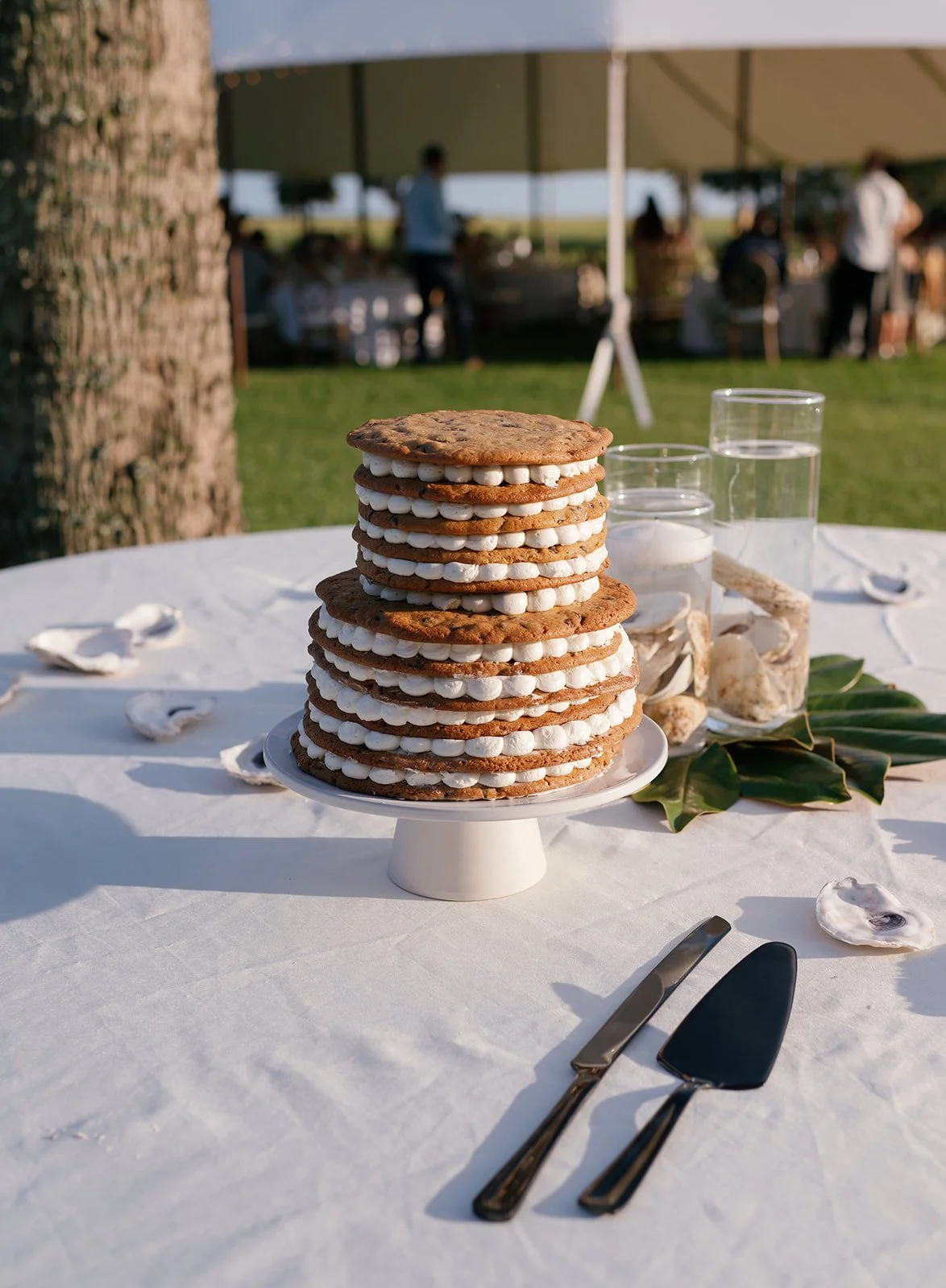 A tall stack of six cookies with white filling on a white cake stand on a table with a white cloth. The table has shell decoration and silverware. Glasses of water and shell decor are in the background under a tent, with people gathered outside.