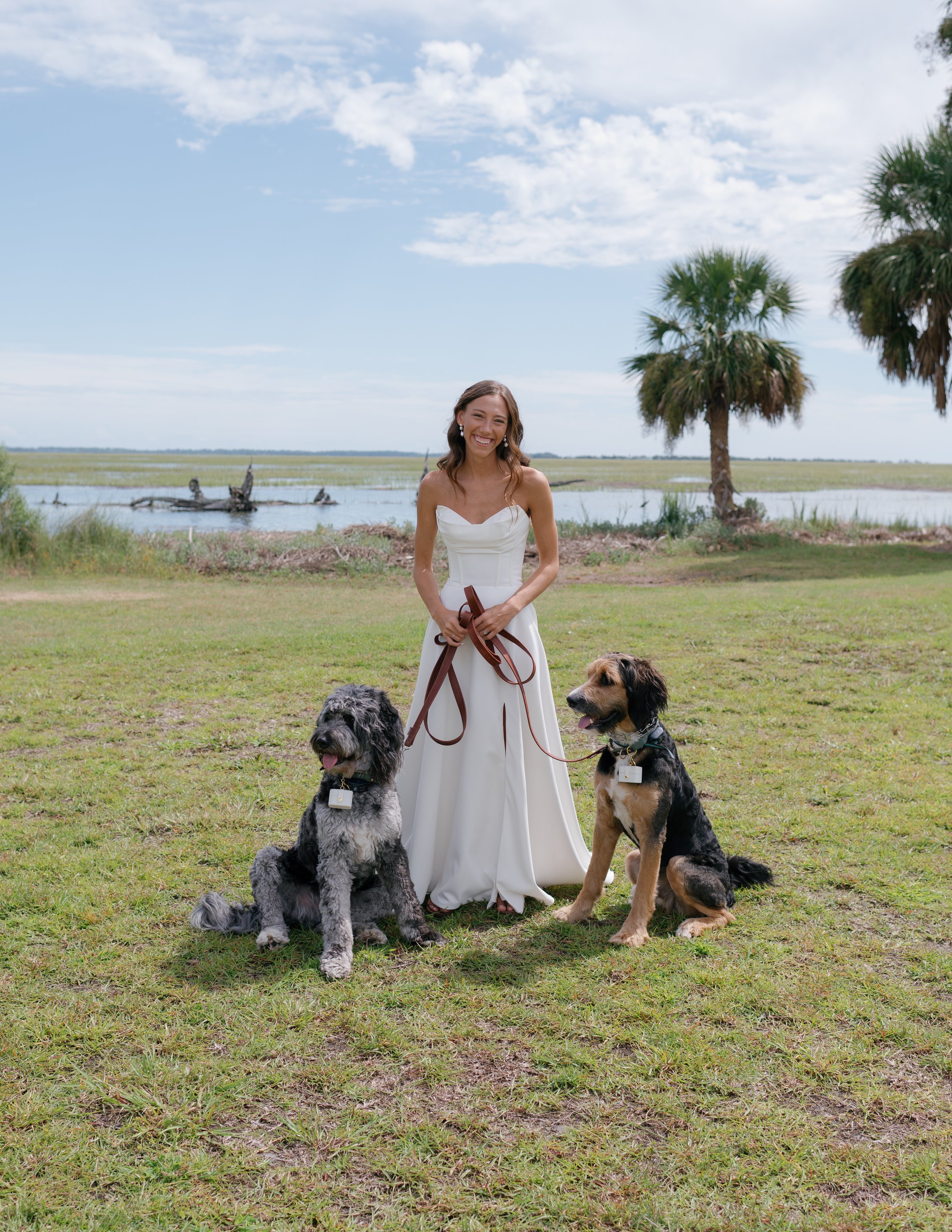 A woman in a white dress stands outdoors on grass by a water body with trees and sky in the background, holding leashes attached to two dogs sitting on her sides.