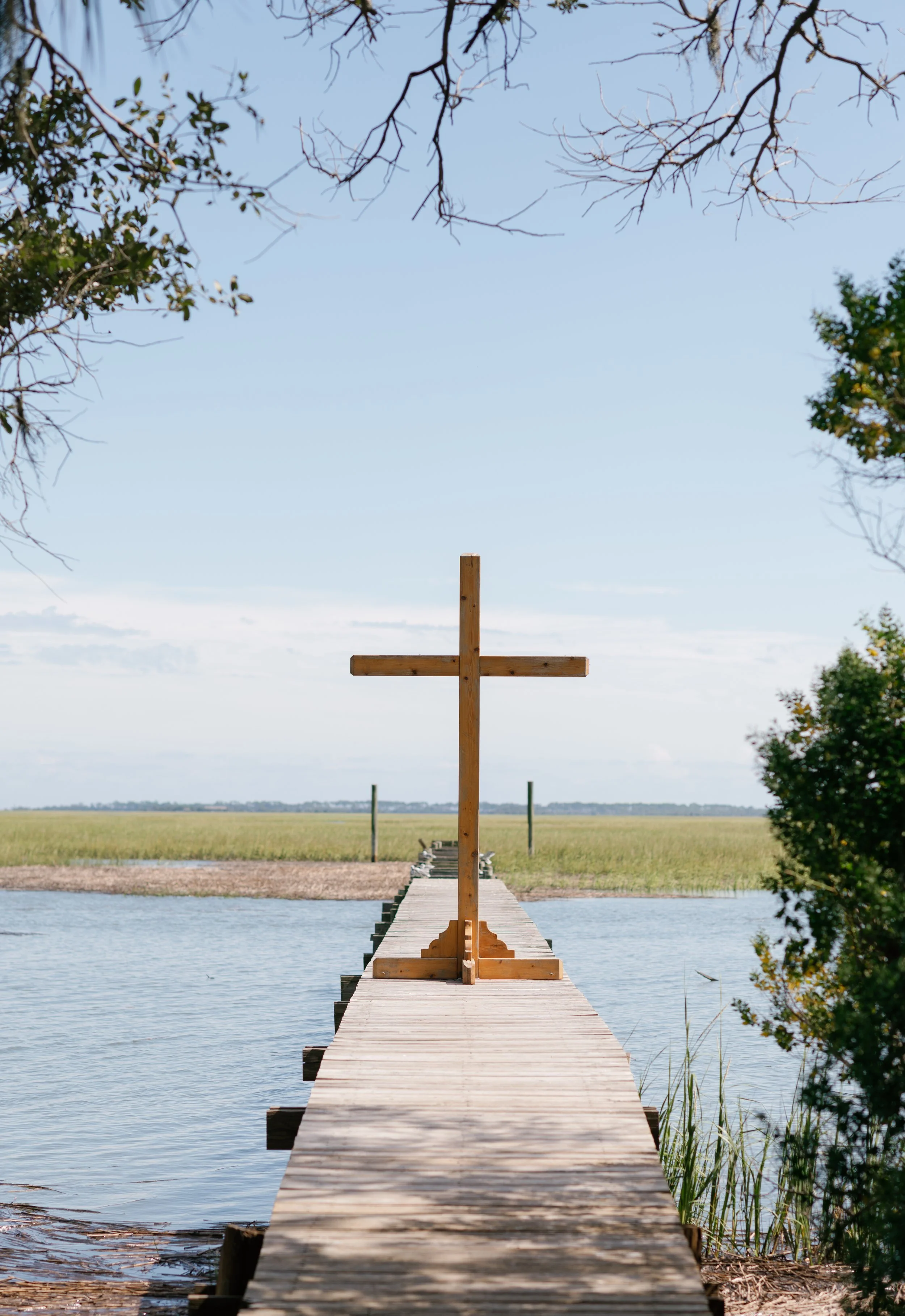 Wooden dock extending into a body of water with a large cross at the end, surrounded by trees and open field in the background.