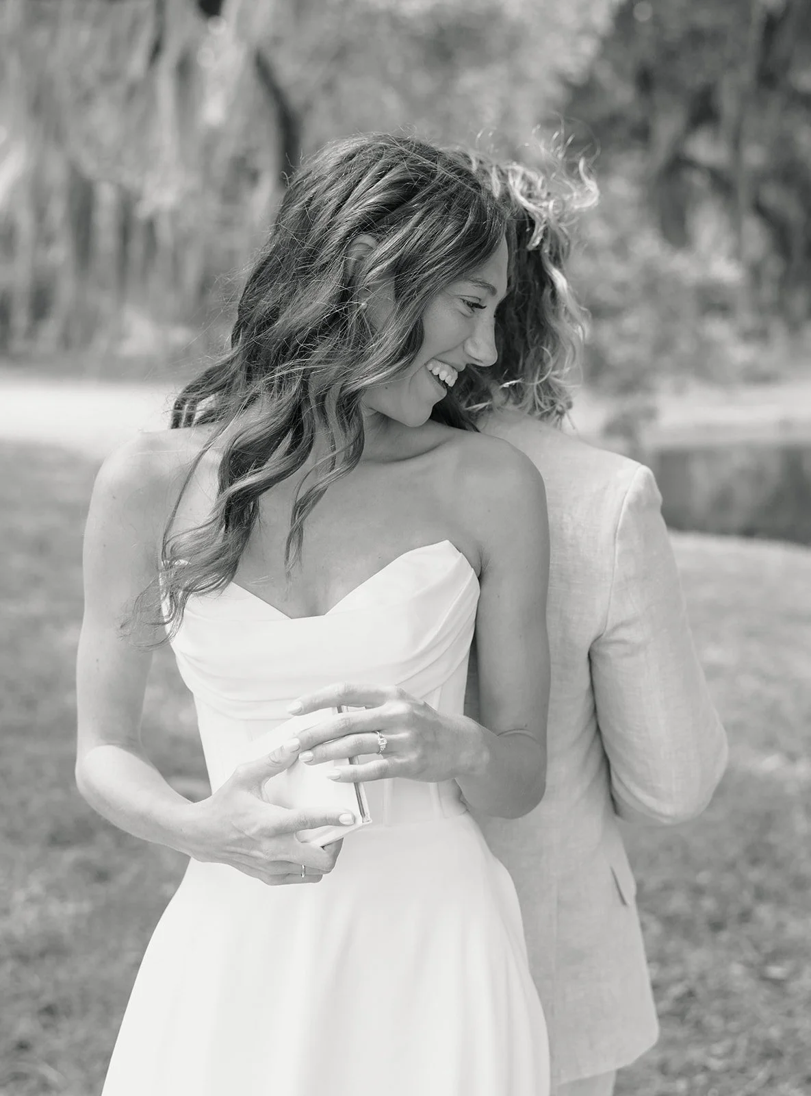 Black and white photo of a woman in a strapless dress smiling and looking down while holding a phone, with a person in a suit standing behind her.