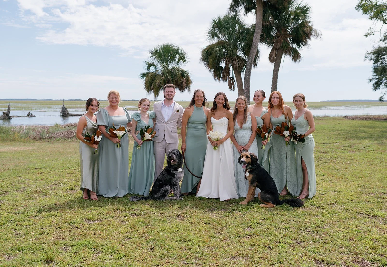 Group of women in pastel dresses and two men in suits, standing outdoors by water with palm trees, holding flowers, with two dogs sitting in front.
