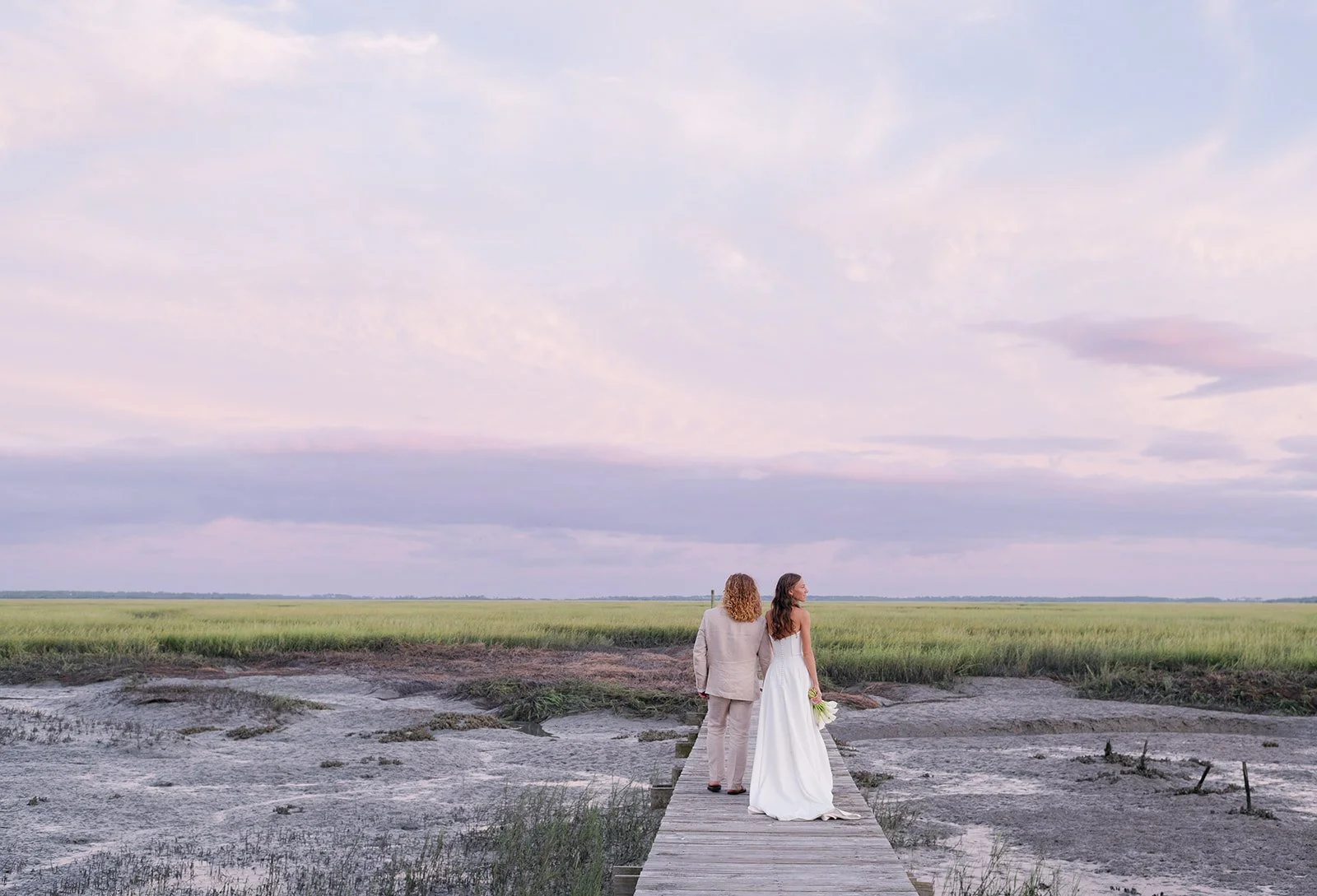 A bride and groom standing on a wooden walkway over a marsh, holding hands and looking at a wide open sky with clouds, during sunset.