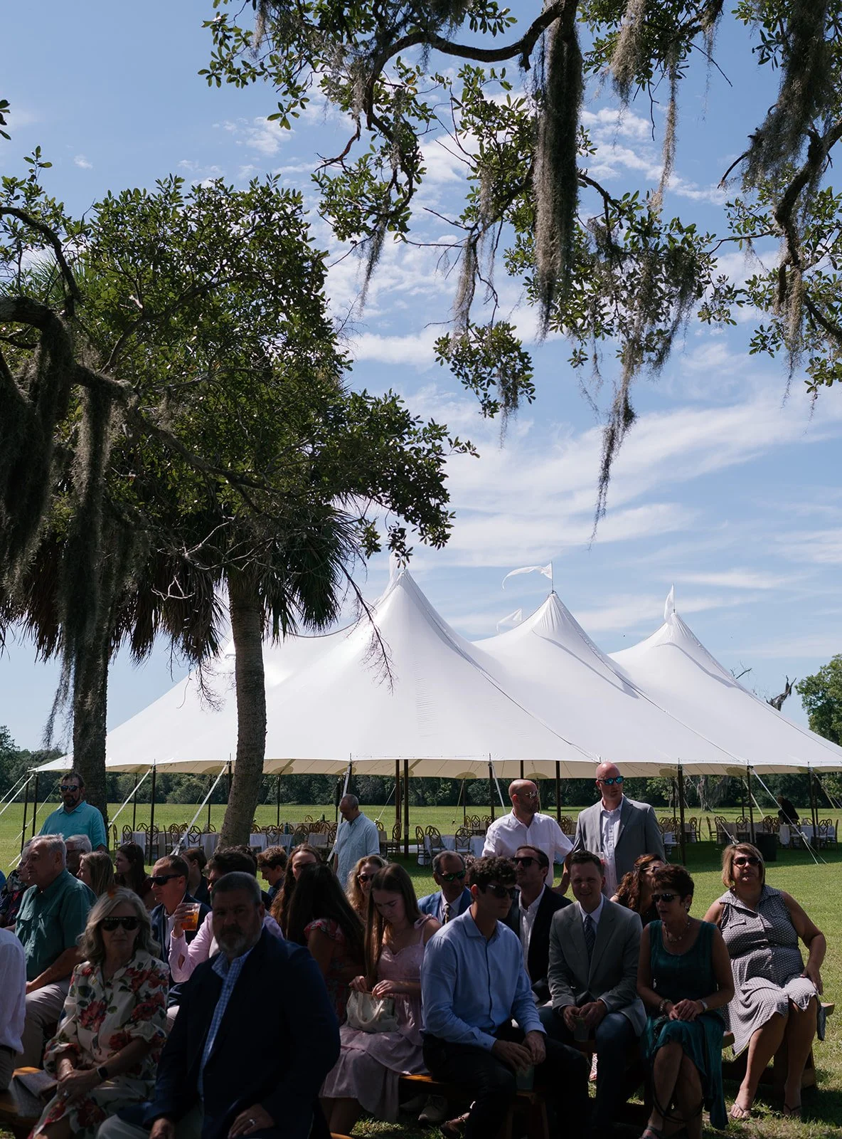 Outdoor gathering of people seated under a large white tent with pointed peaks, surrounded by trees with hanging moss, on a sunny day with a blue sky and scattered clouds.