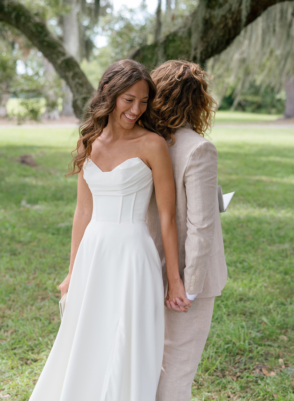 Two women in wedding attire standing outdoors on a lush green lawn, holding hands and smiling with their heads close together, surrounded by trees.