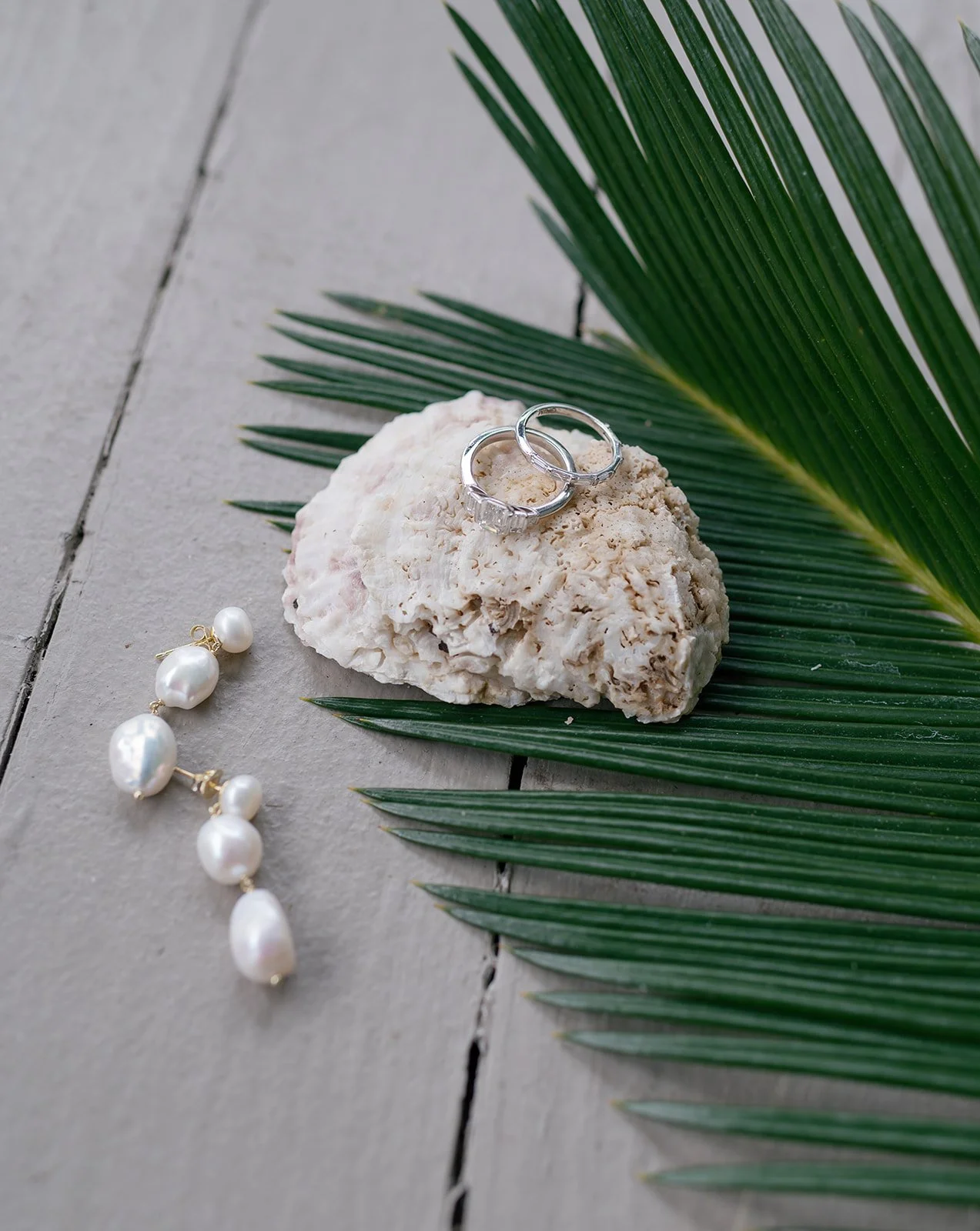 Silver rings on a coral rock, pearl earrings, and a pearl necklace with tropical palm leaves on a gray wooden surface