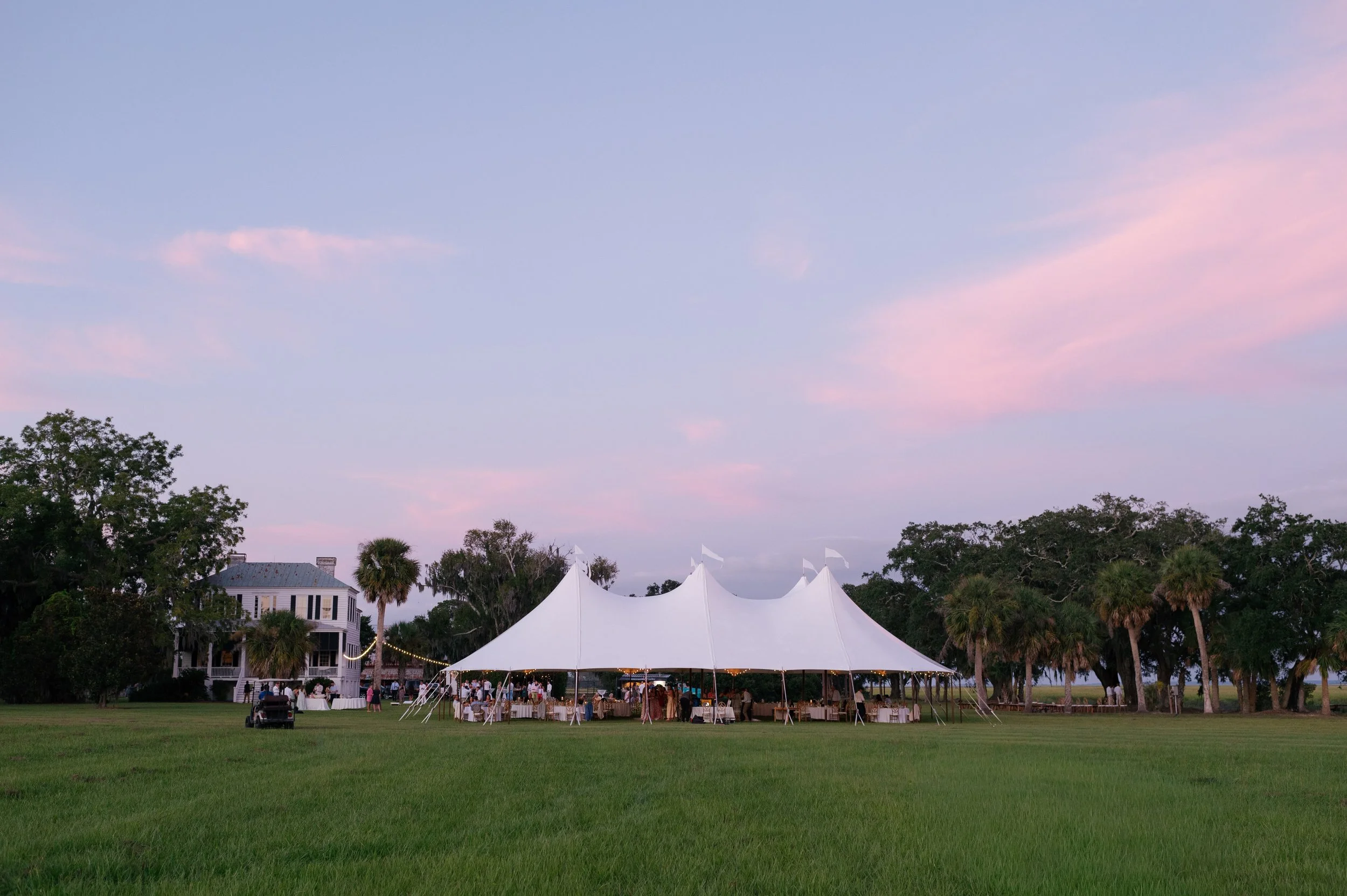 Outdoor event setup with a large white tent, tables, and chairs on a grassy field surrounded by trees, under a pastel-colored sky at dusk.