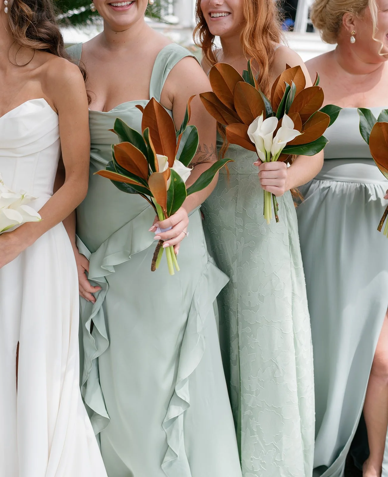 Group of women dressed in pastel dresses holding bouquets of white calla lilies and brown-tinted leaves at a wedding.