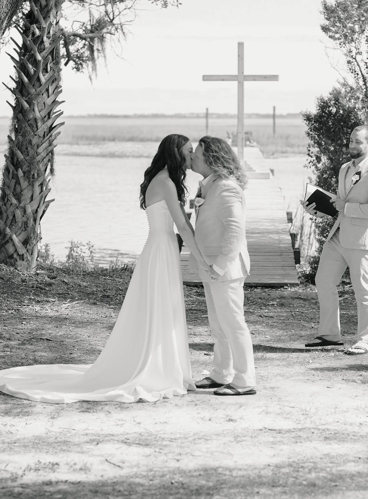 A black and white photo of a wedding ceremony where a bride and groom are kissing on a dock by the water, with a priest or officiant holding a book to the right, and a cross in the background.