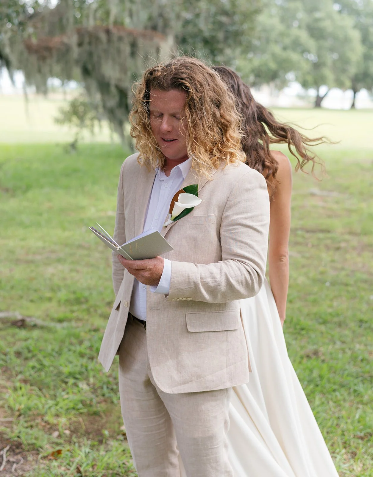 A man reading a card in a beige suit at an outdoor wedding, with a woman in a white dress behind him, in a grassy area with trees.
