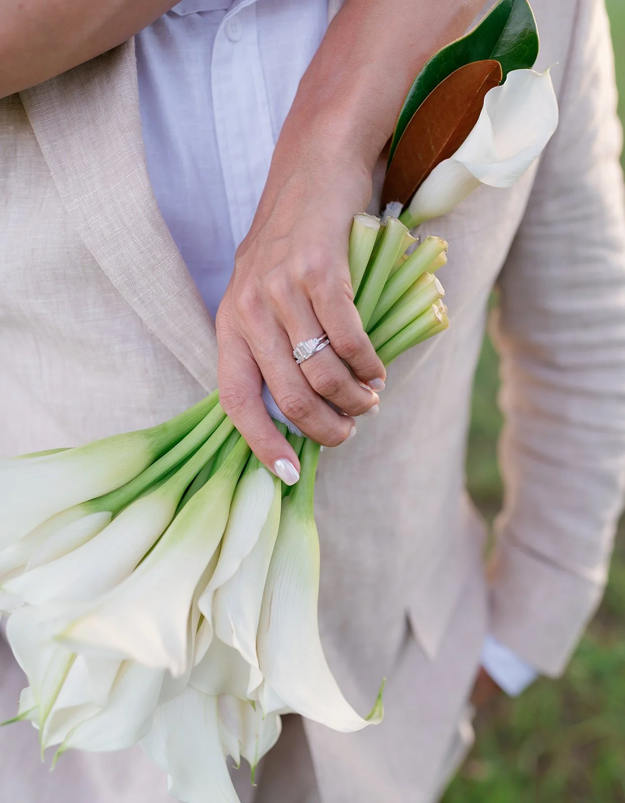 A person wearing a light-colored blazer and white shirt holds a bouquet of white calla lilies and green leaves, with a wedding ring visible on their finger.