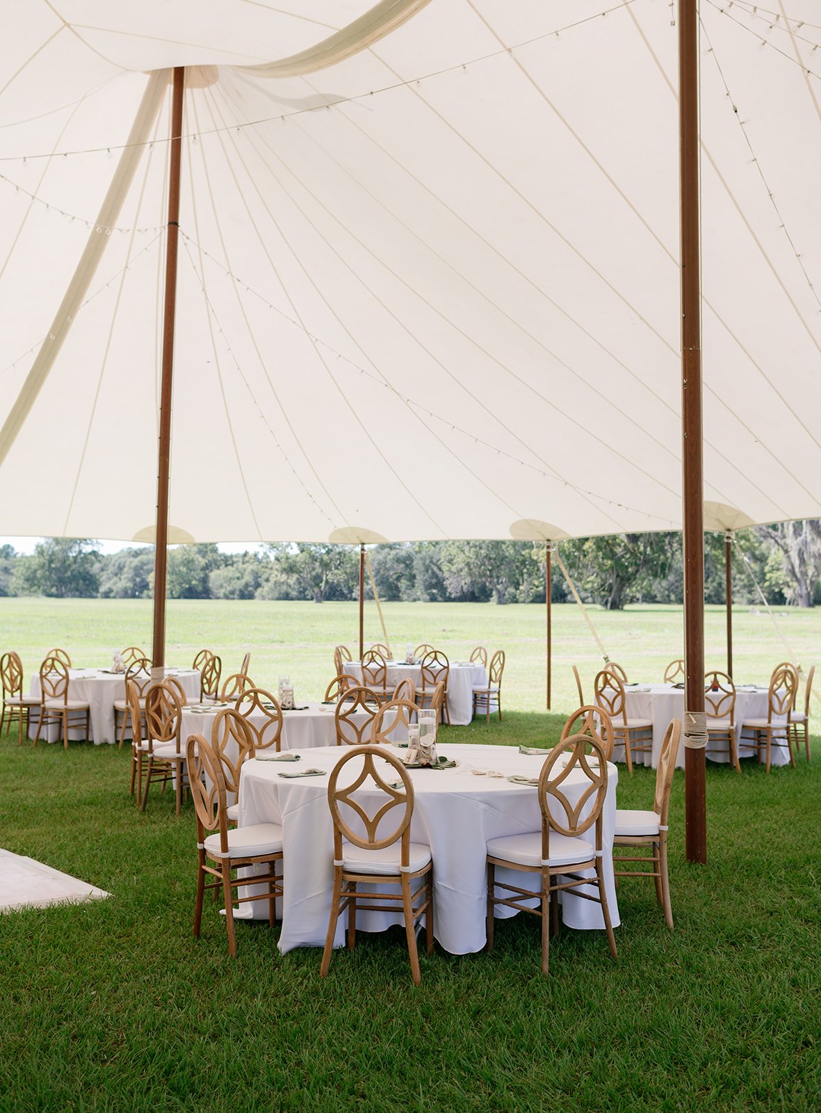 Outdoor event setup with round tables covered with white tablecloths, surrounded by wooden chairs, under a large white canopy tent on a grassy area with trees in the background.
