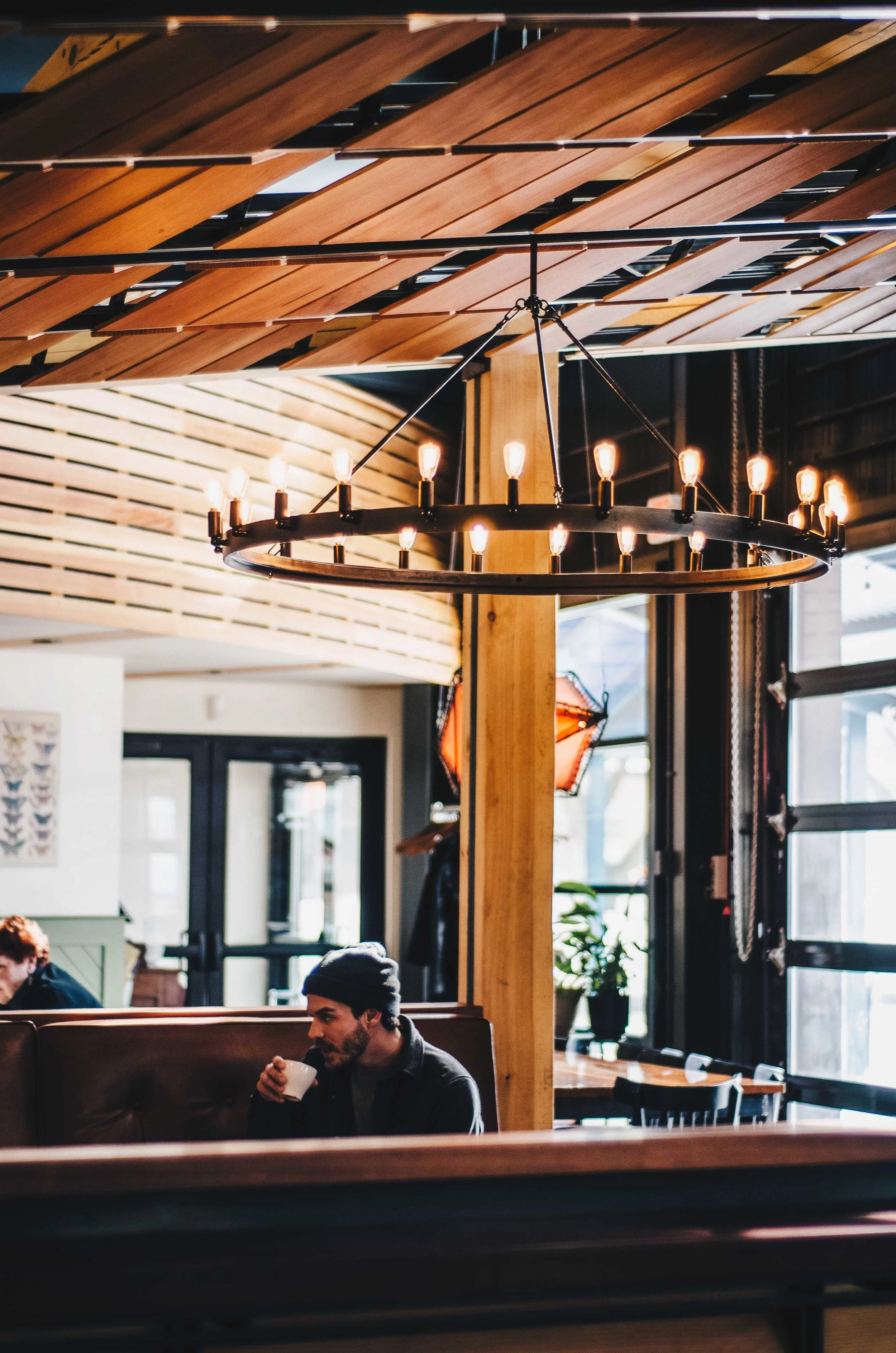 The image shows a modern indoor space with a wooden ceiling and a circular chandelier with exposed light bulbs hanging from the ceiling. There are large windows allowing natural light to enter and a person sitting at a table drinking from a cup, wearing a dark beanie and jacket.