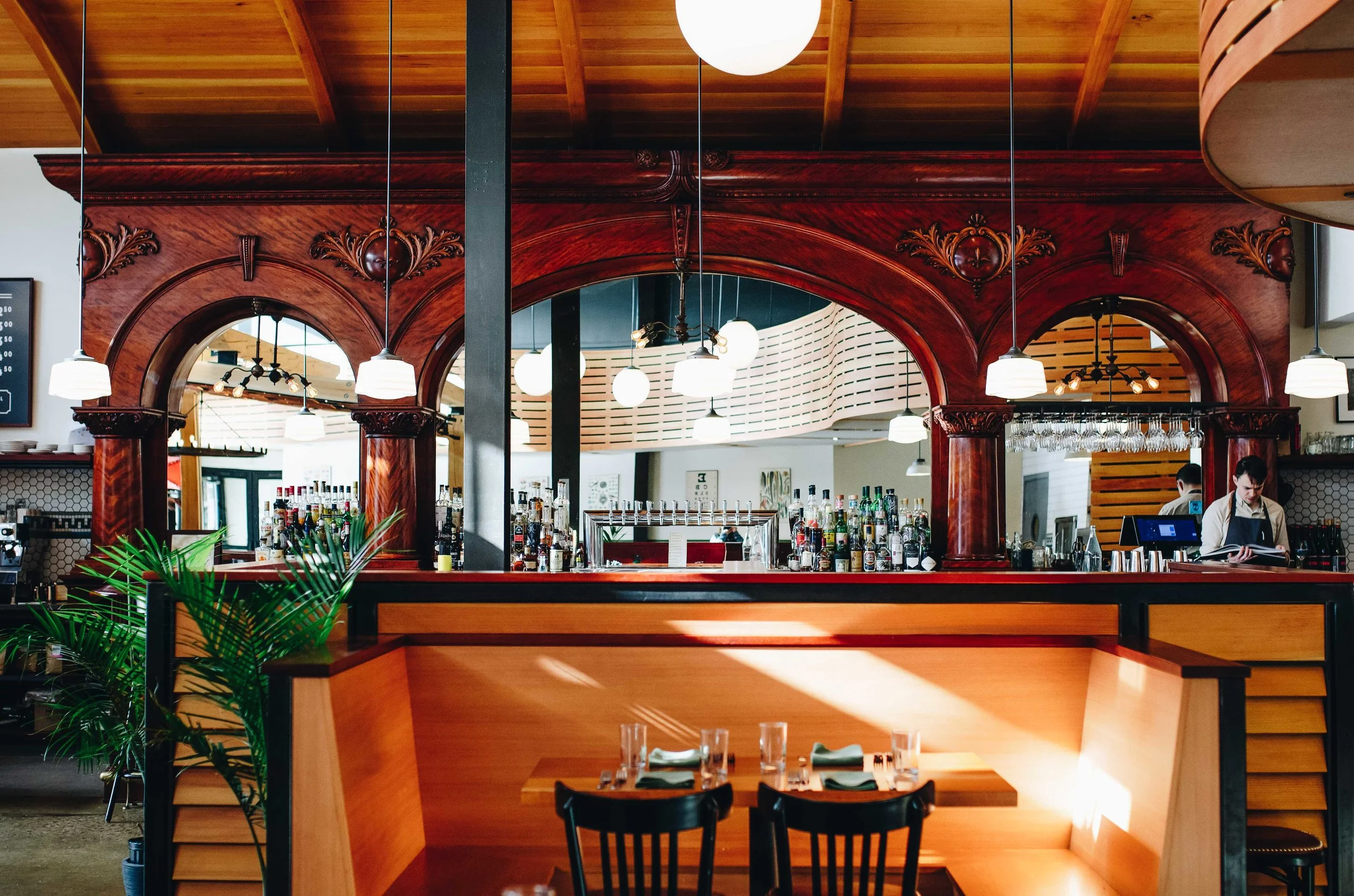 Interior of a restaurant with wood accents, hanging white pendant lights, bar area with bottles, and tables set with glasses and napkins.