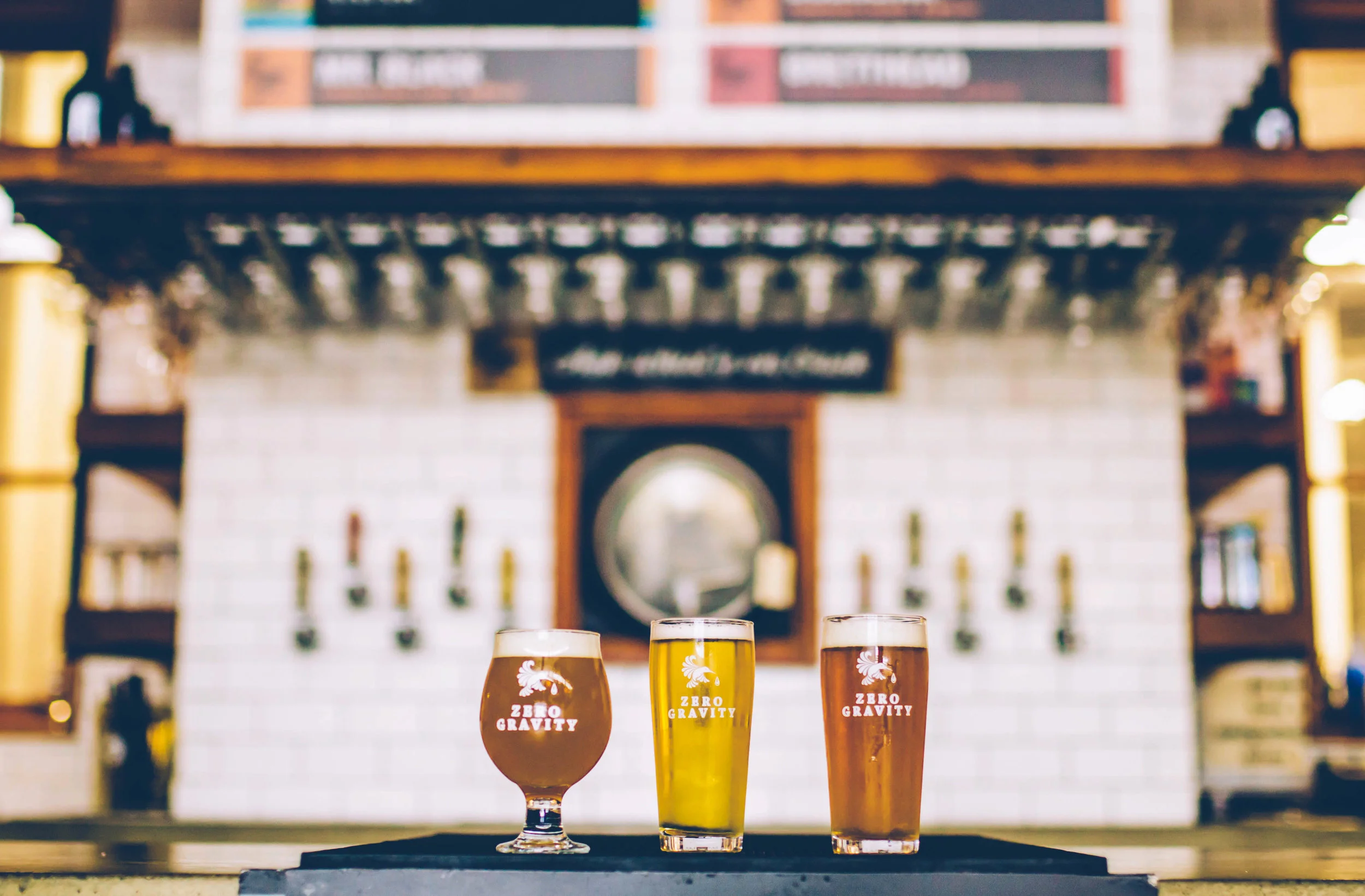 Three beer glasses in a row on a table with a bar or pub background. The glasses display the logo 'Zero Gravity' on each glass.