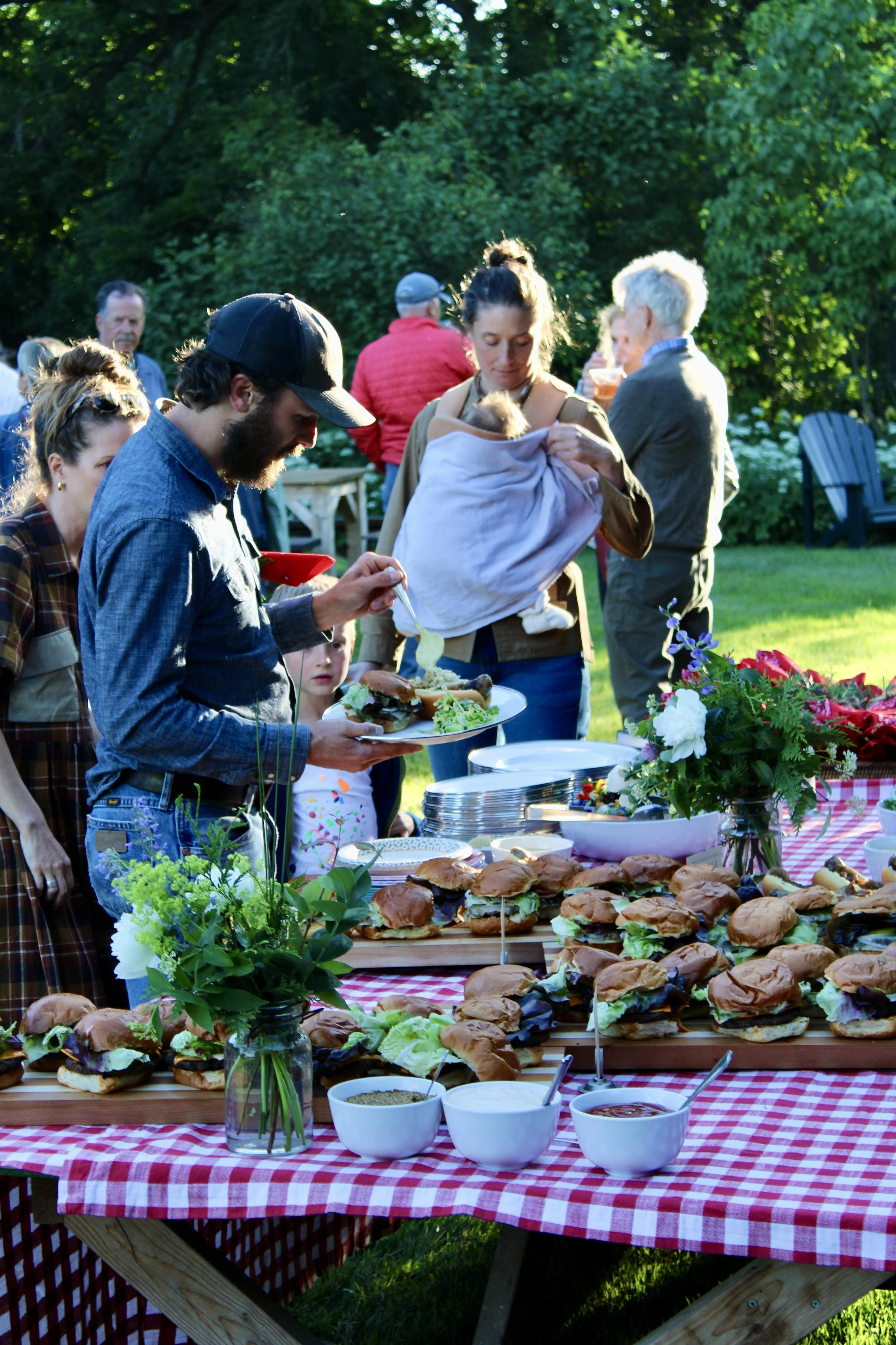 People gathered around a table outdoors, enjoying a barbecue with sandwiches, salads, and drinks, during a sunny day in a park.
