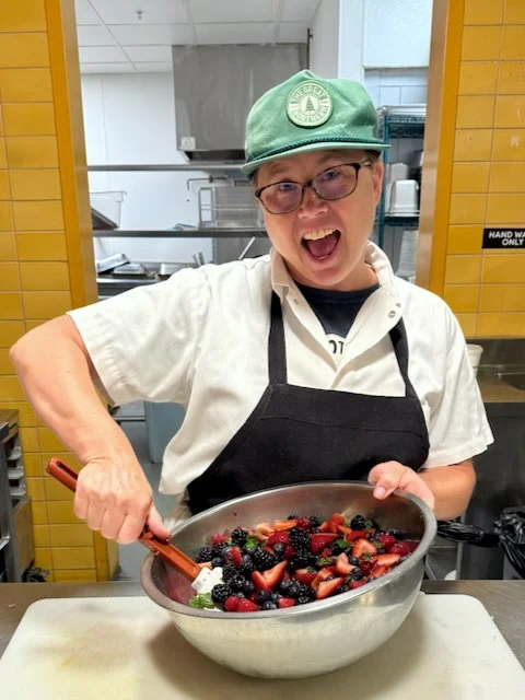 Person in a white uniform and green cap holding a large bowl of mixed berries, smiling excitedly in a kitchen setup.