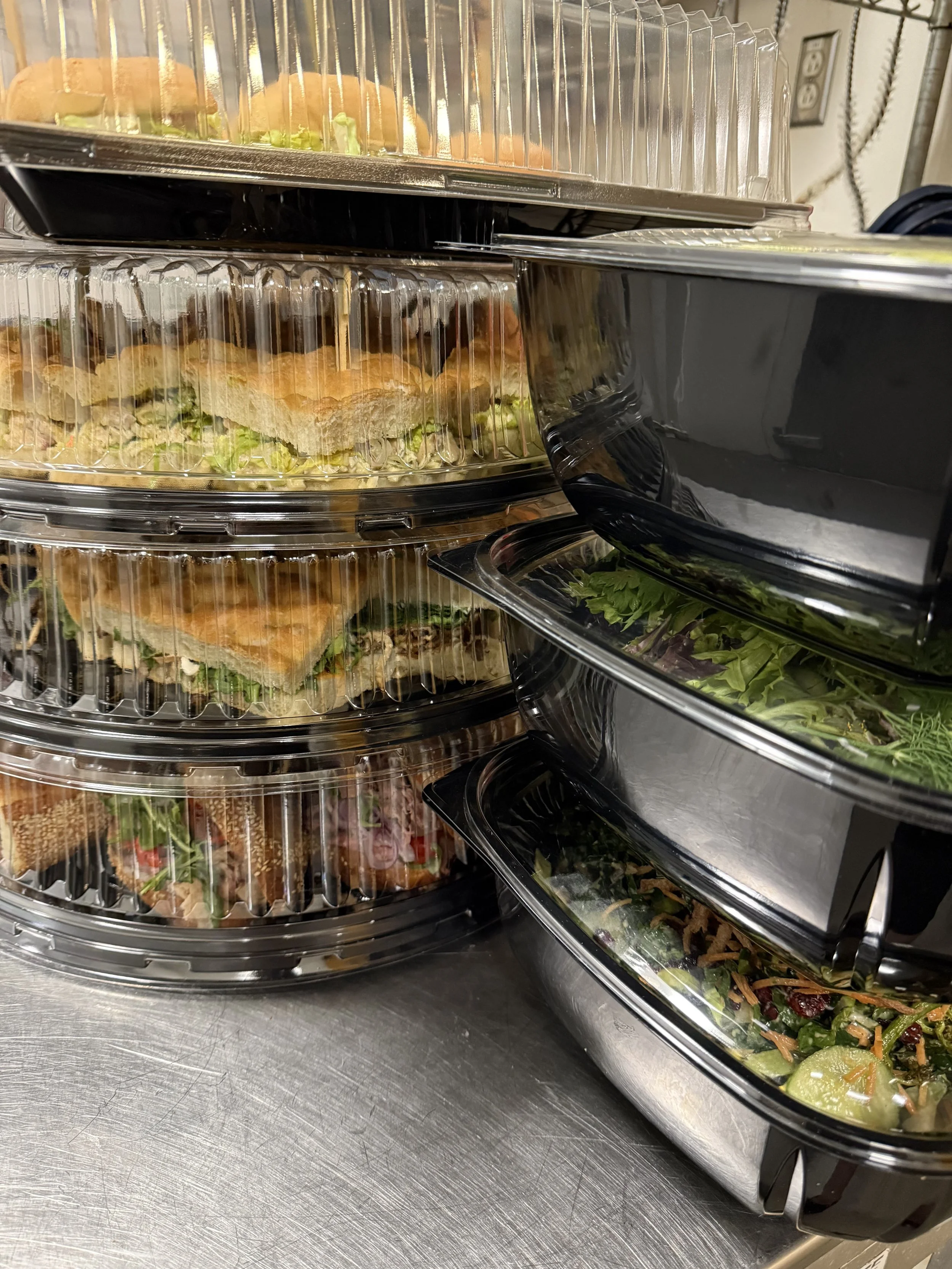 Stacked black plastic food containers filled with salad, sandwiches, and wraps, placed on a stainless steel counter.