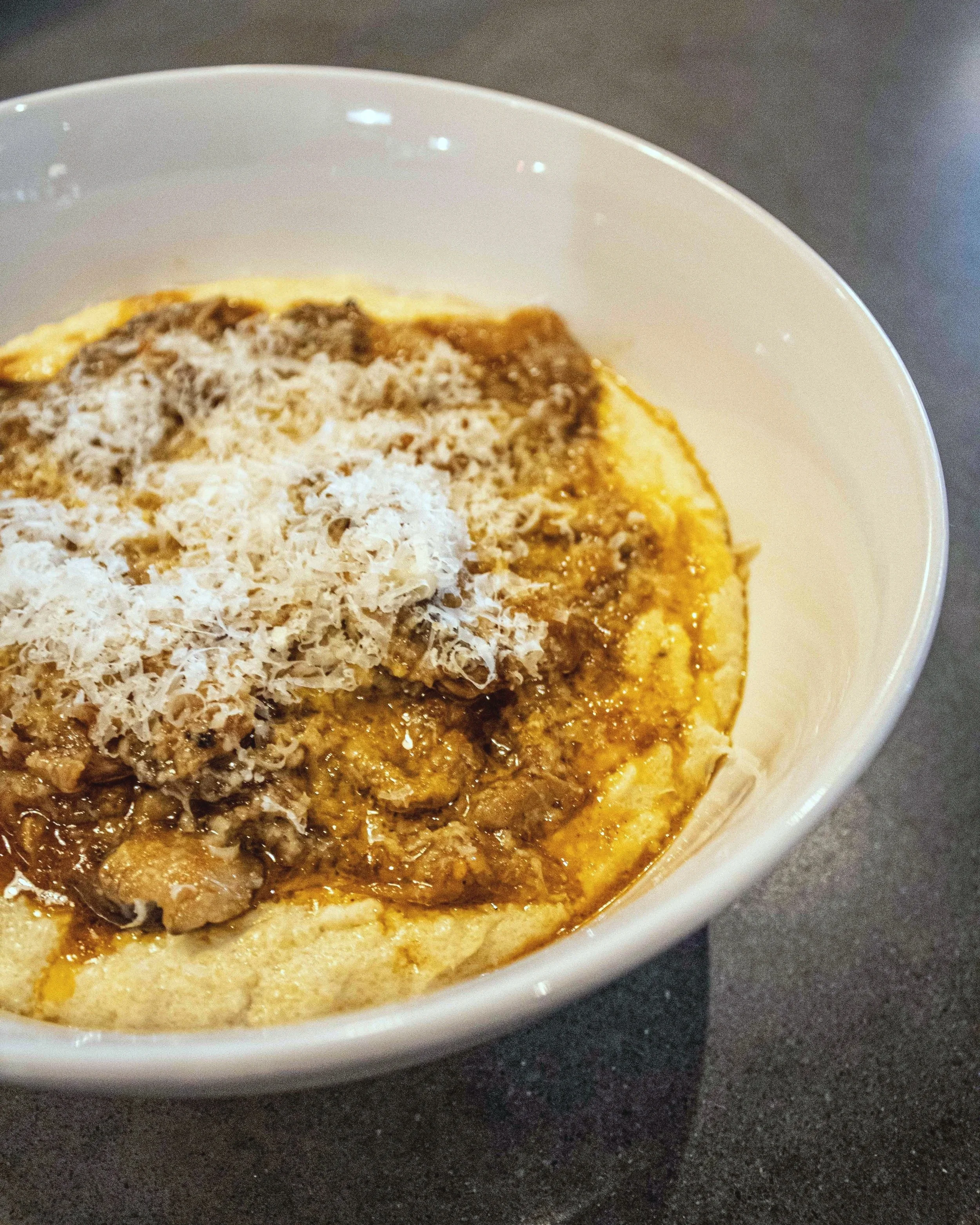 Close-up of a bowl with a creamy dish topped with shredded cheese, ground meat sauce, and herbs or spices on a dark surface.