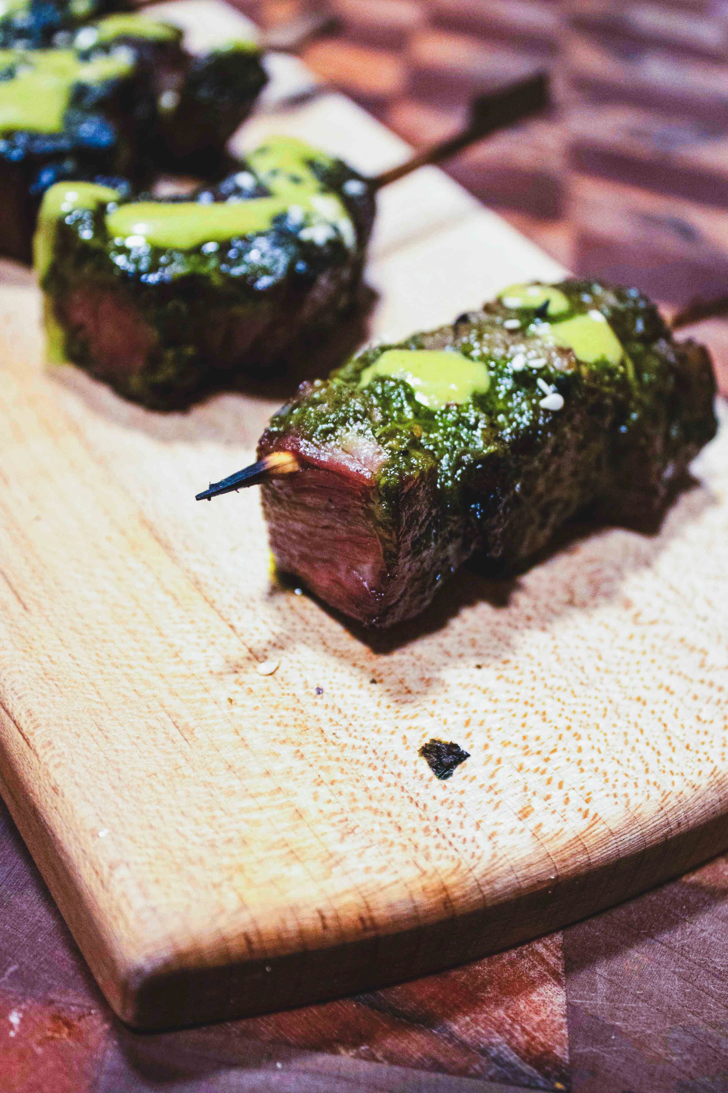 Close-up of two slices of beef topped with green sauce and sesame seeds on a wooden cutting board.