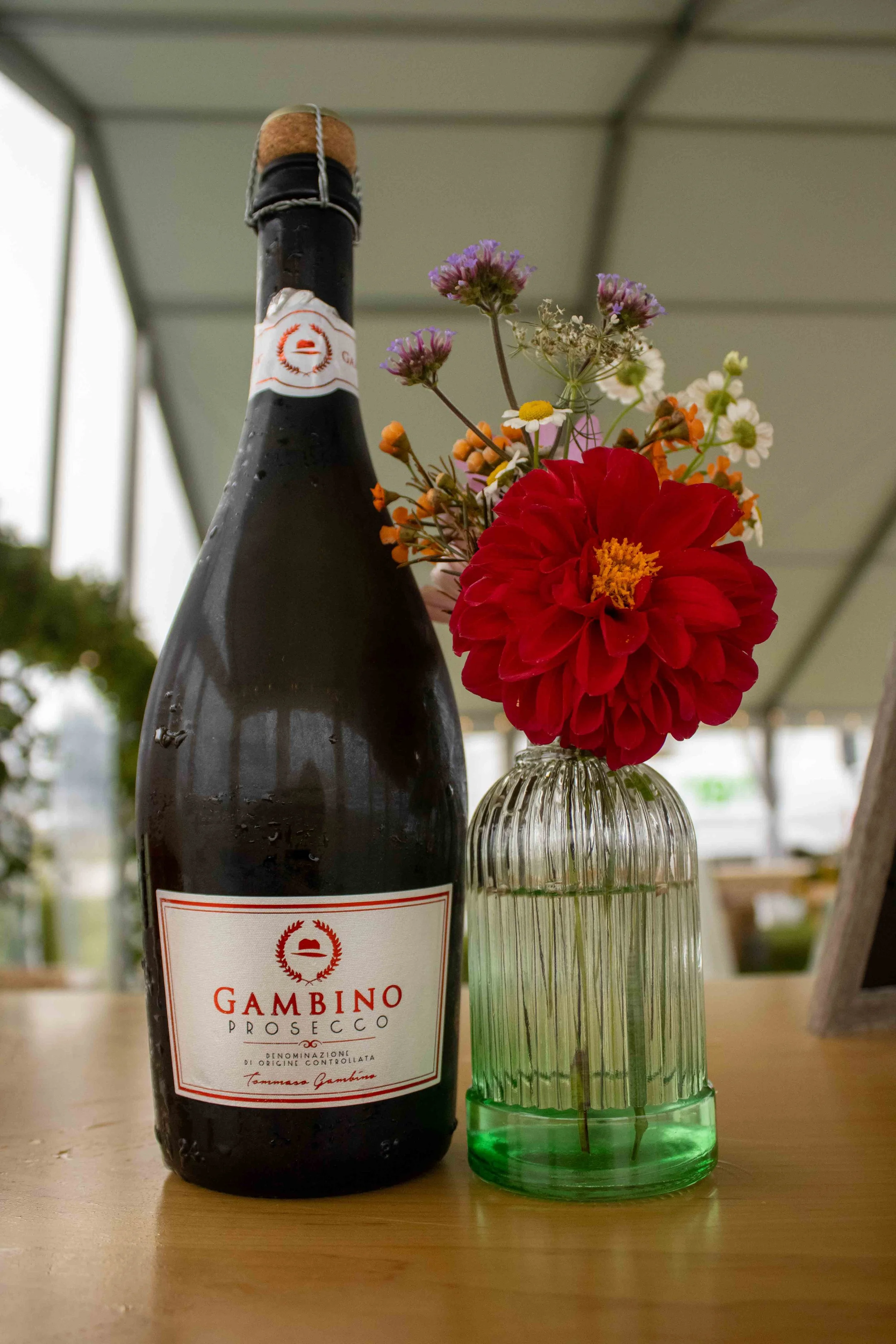 A bottle of Gambino Prosecco with a bouquet of colorful flowers in a glass vase, placed on a wooden surface, with an outdoor setting in the background.