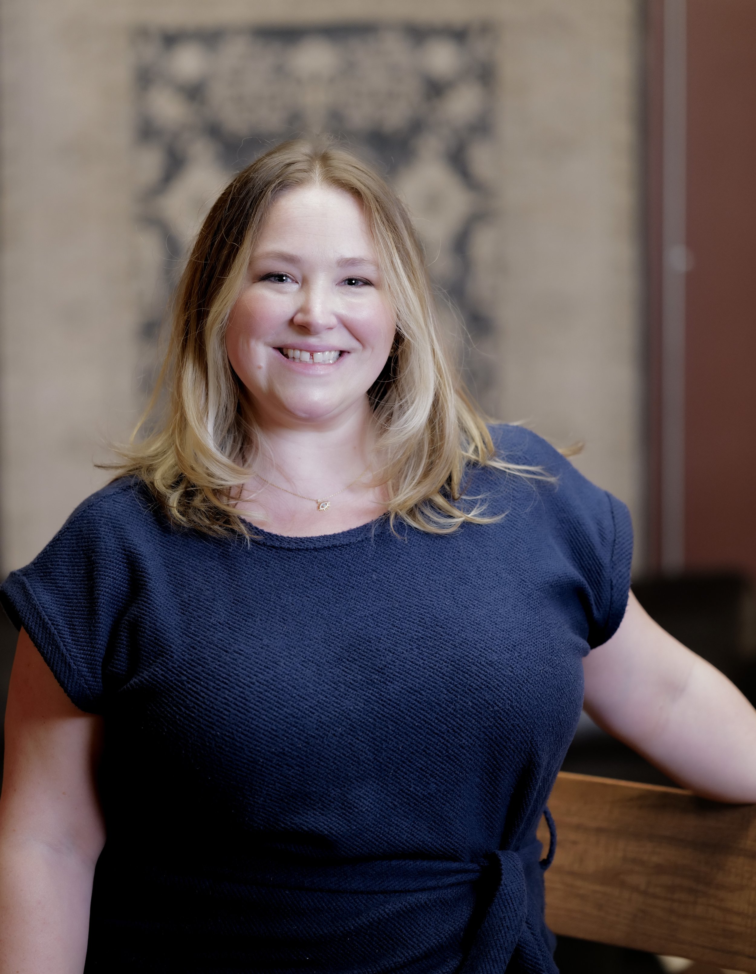 A woman with blonde hair smiling in a navy blue top, seated at a wooden table against a brick wall with a decorative metal object in the background.