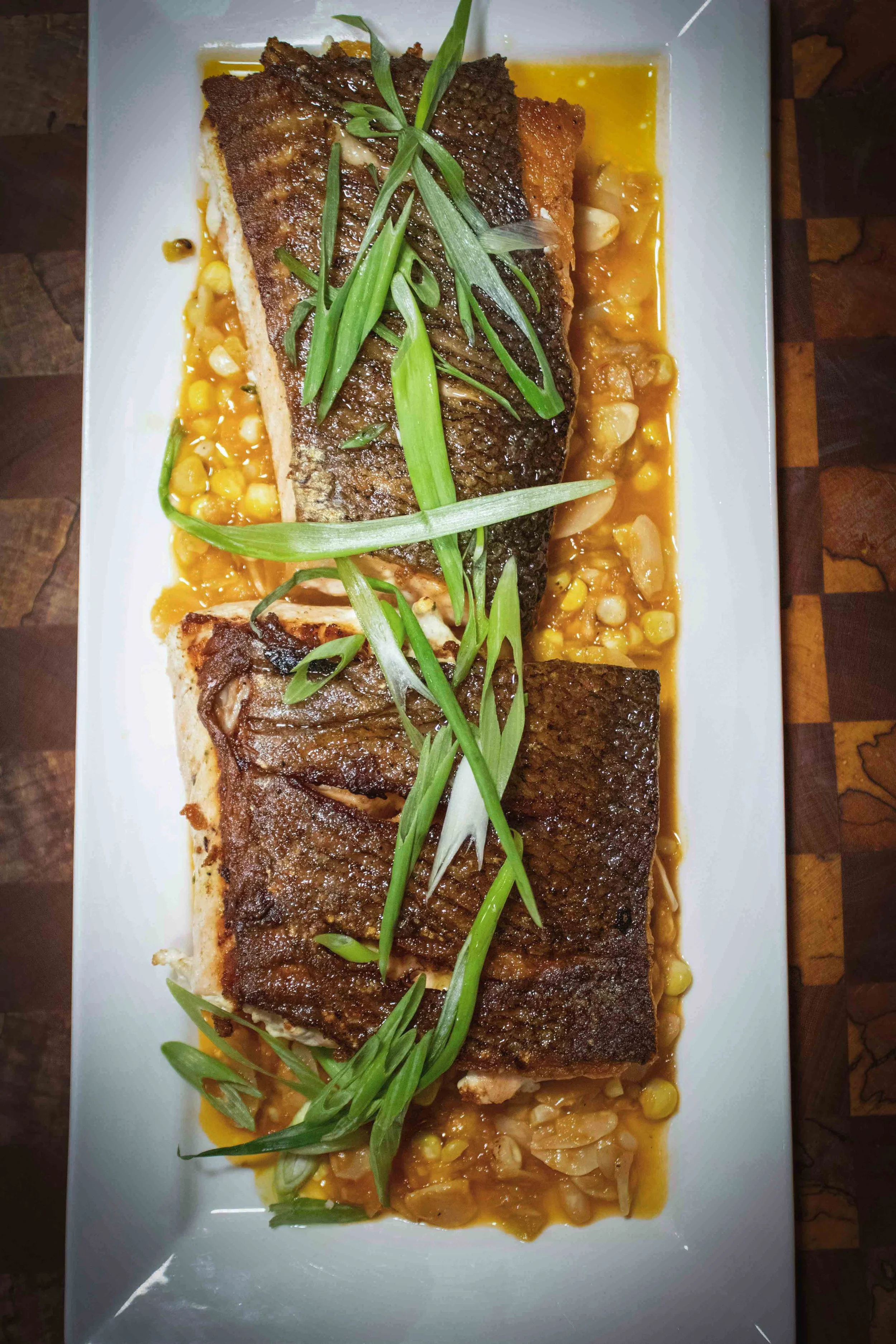 A dish consisting of grilled fish fillets topped with green onions, served on a white rectangular plate with corn and sauce underneath.