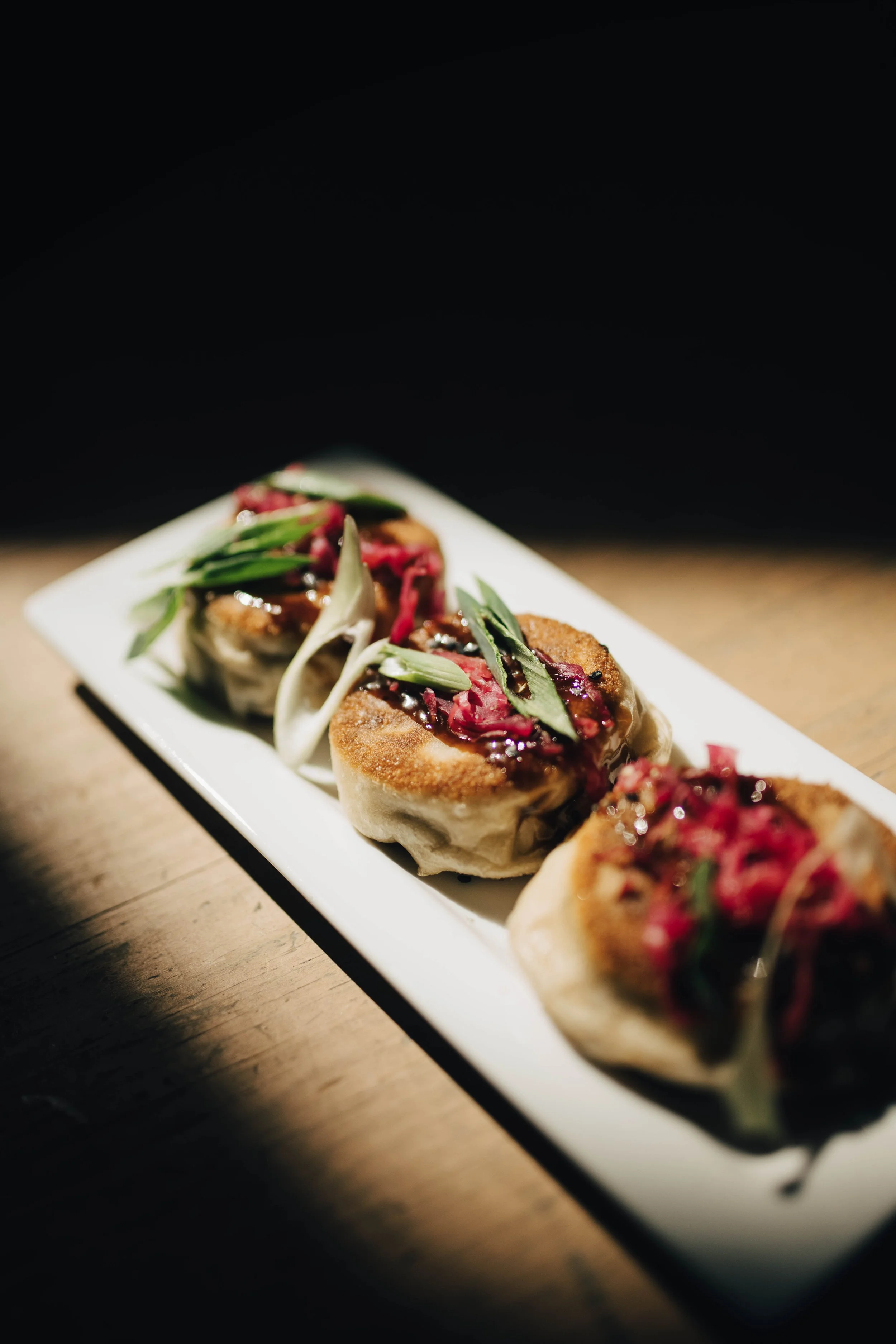 Close-up of several gourmet appetizers on a white rectangular plate, garnished with herbs and topped with a dark sauce, on a wooden table with a dark background.
