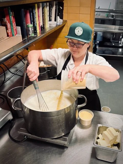 A person wearing glasses, a green baseball cap, and a white chef's coat, stirring a large pot on a stovetop in a kitchen. They are pouring a yellow liquid from a cup into the pot, with a whisk in their other hand. The kitchen has shelves with books and ingredients, and there is a small bowl and a tin with blocks of butter on the counter.