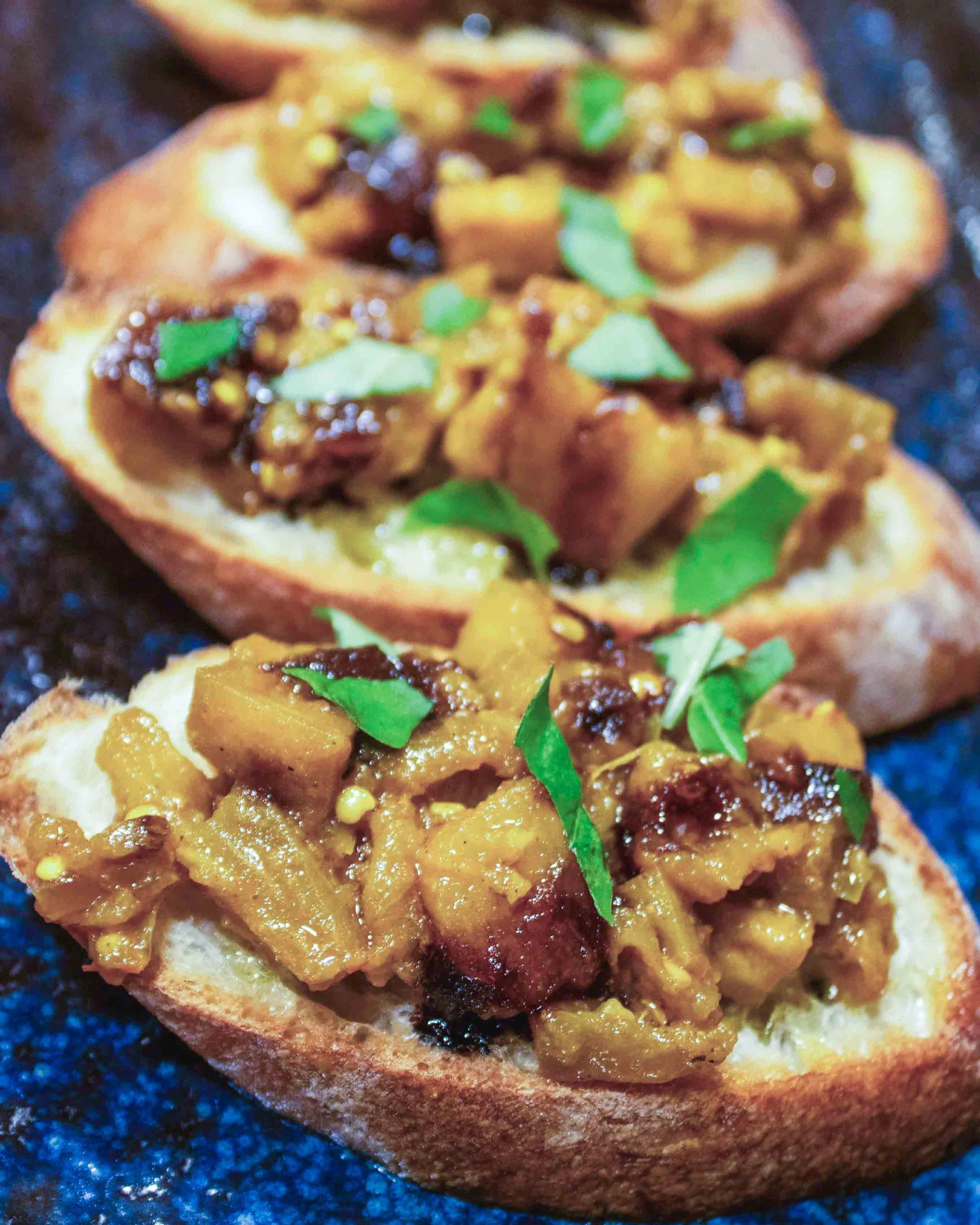 Close-up of a piece of bread with toppings including mango chutney, diced mango, fresh herbs, and a dark sauce.