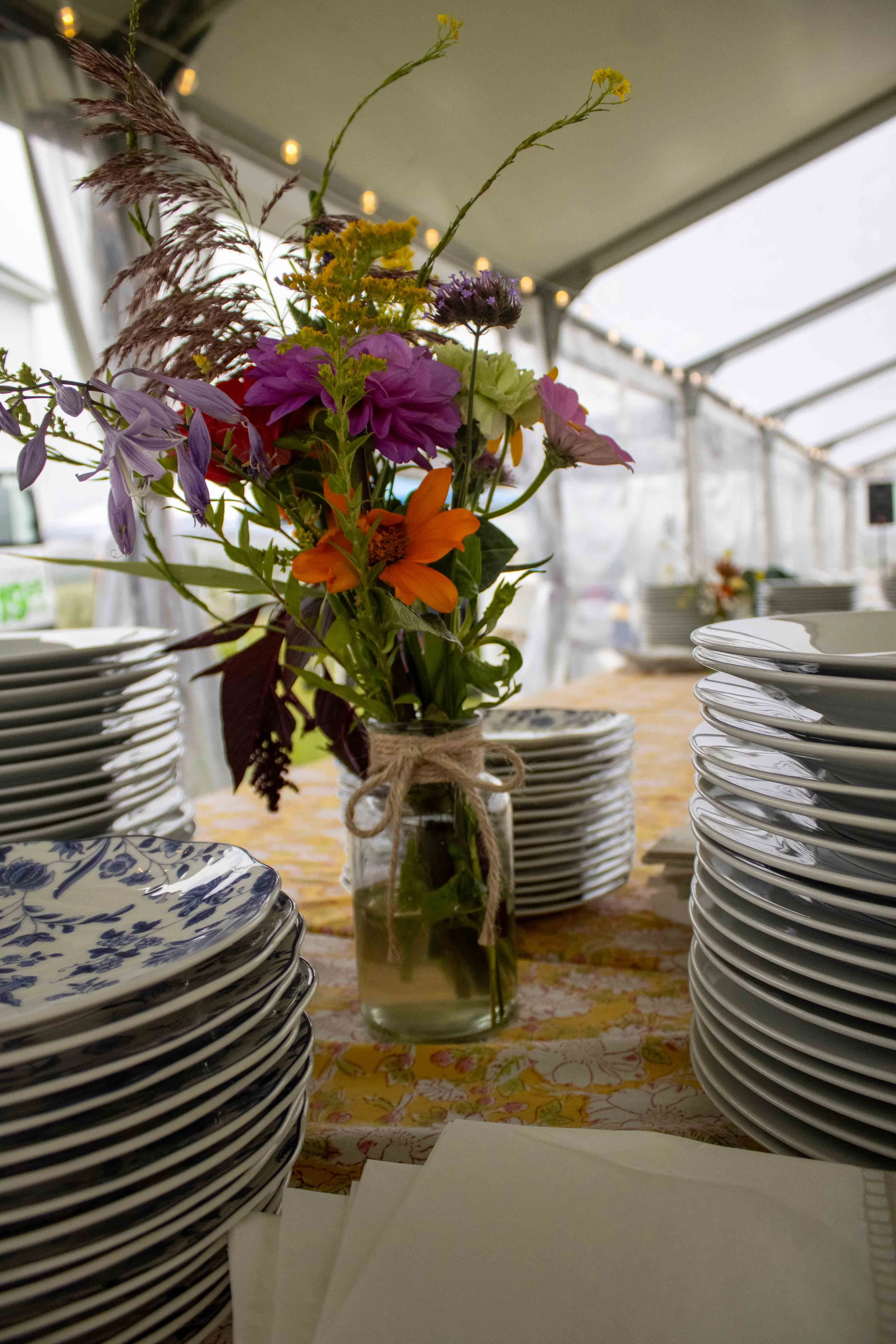A floral centerpiece in a glass jar with a twine bow, surrounded by stacks of white ceramic plates on a decorated table inside a tent with string lights.
