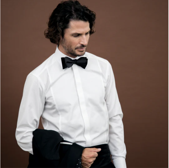 Man with curly hair wearing a white dress shirt and black bow tie, holding a jacket, against a brown background.