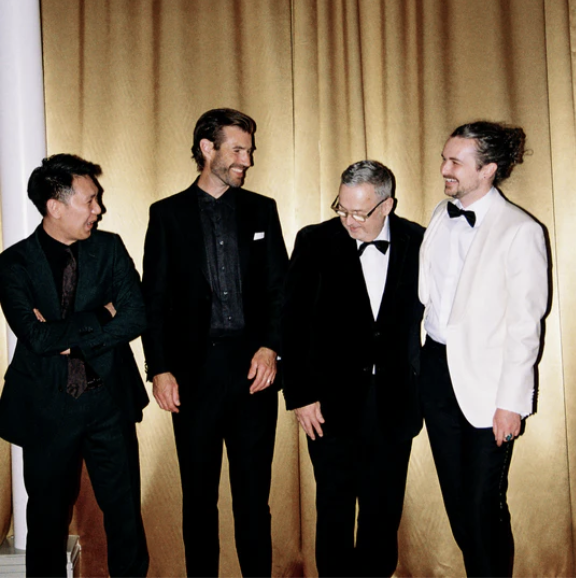 Four men in formal attire, standing and smiling in front of beige curtains.