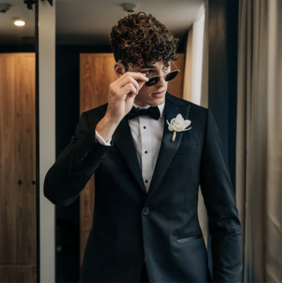 A young man with curly hair in a tuxedo, wearing sunglasses and holding them with one hand, standing indoors near a window with curtains.