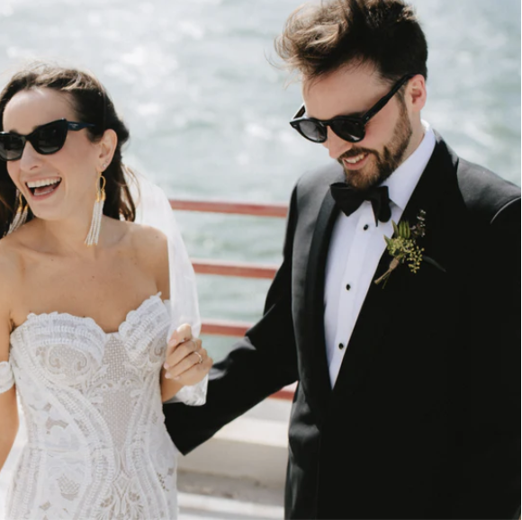A smiling bride wearing sunglasses, a white lace wedding dress, and gold earrings, standing next to a groom in a black tuxedo with a bow tie and sunglasses, outdoors near water during daytime.