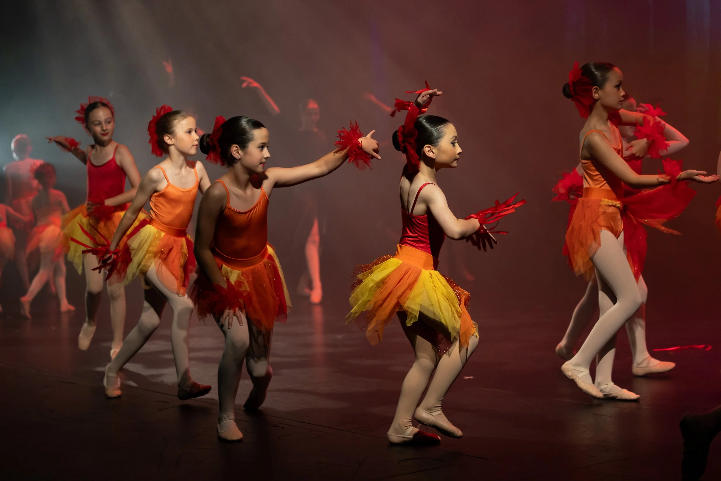 Group of young girls dressed in colorful ballet costumes performing on stage with red and orange tutus, some with headbands or bows, under stage lighting.