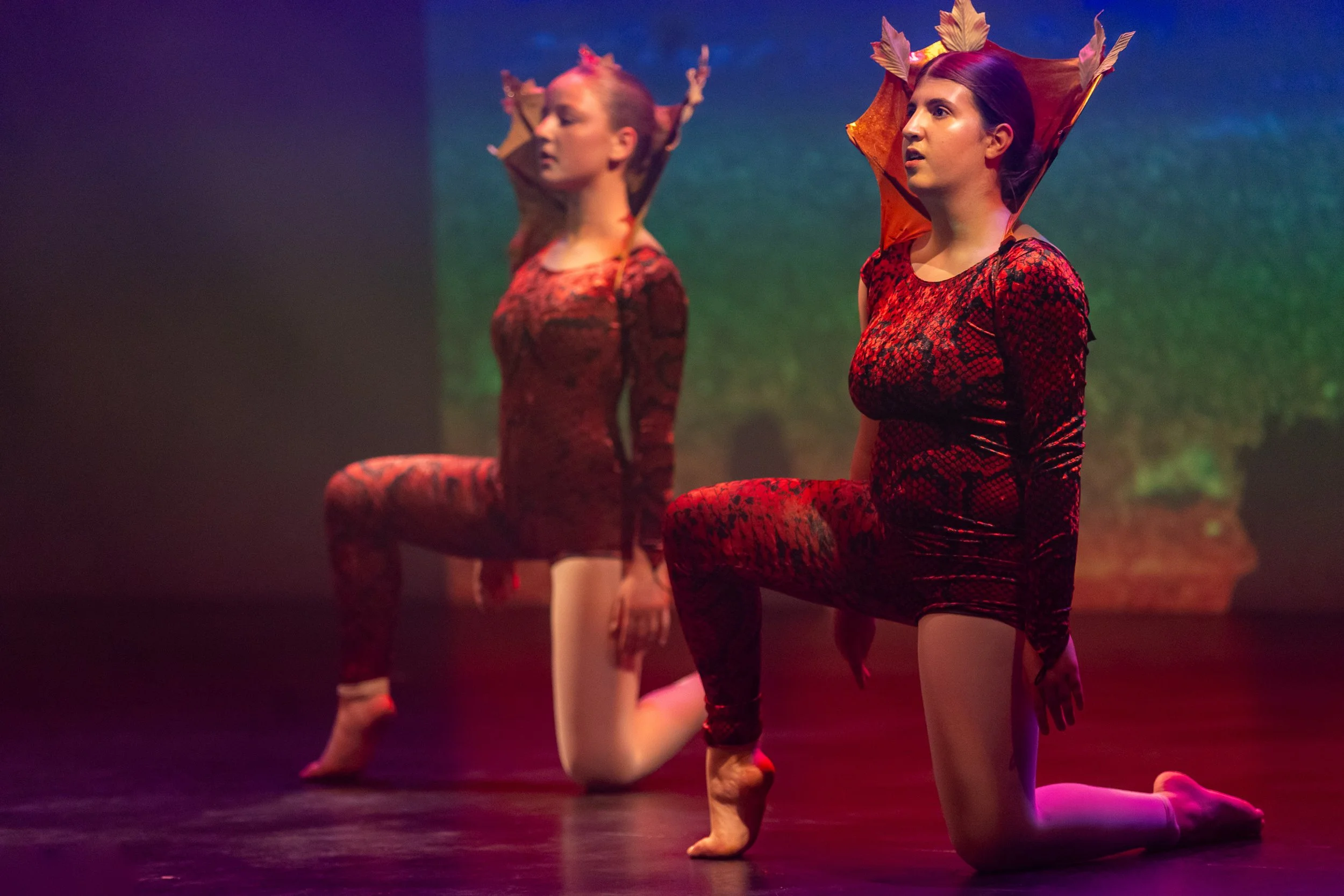 Two women dressed in red, black, and orange costumes with headpieces resembling leaves, performing a dance on stage with a nature-themed backdrop.