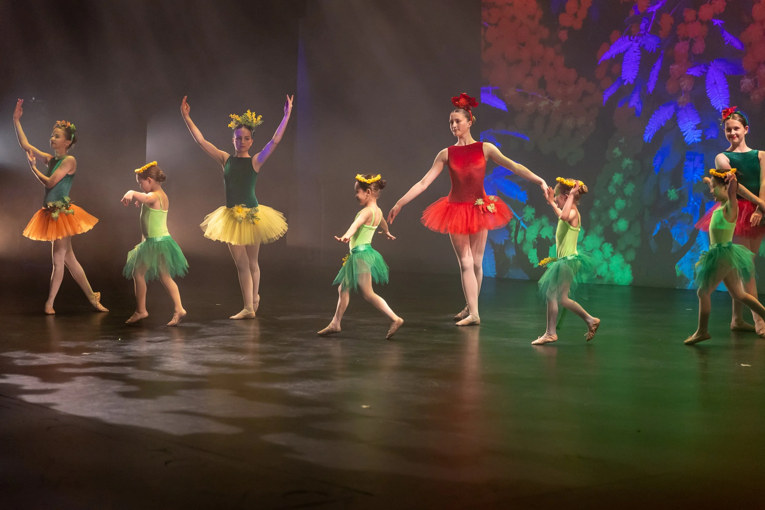 Group of young ballet dancers dressed in colorful costumes performing on stage with a nature-themed backdrop.
